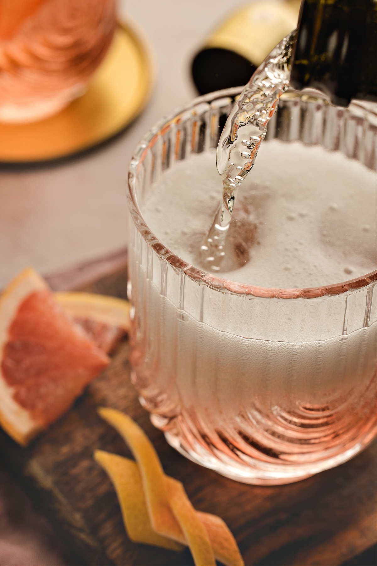 Sparkling wine being poured into a glass of Lillet Rosé spritz, creating a foamy layer over the pale pink drink with grapefruit slices and twists nearby.