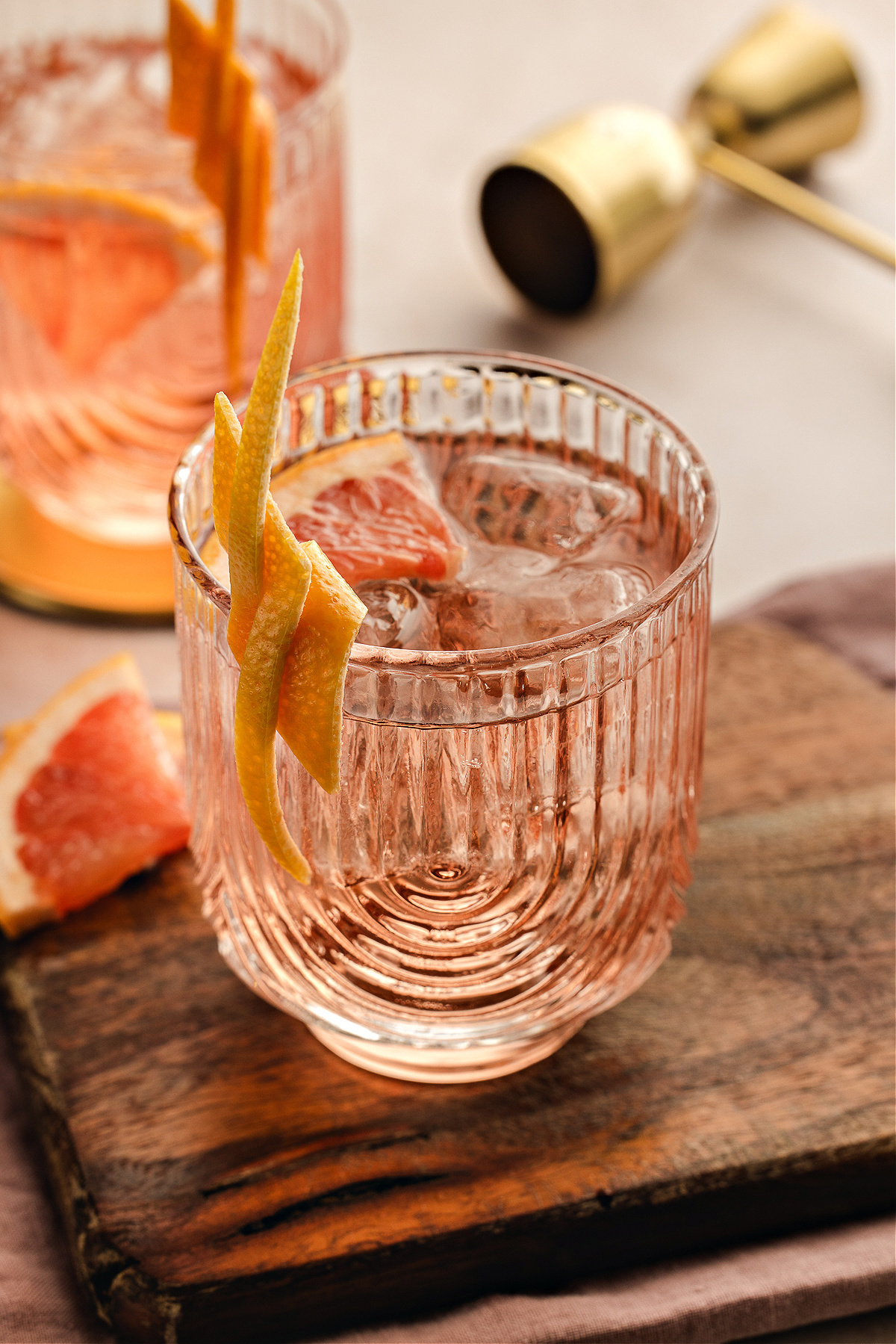 A pale pink Lillet Rosé spritz served over ice in a textured glass, finished with a long grapefruit twist and a slice tucked inside, with bar tools and another spritz blurred in the background.