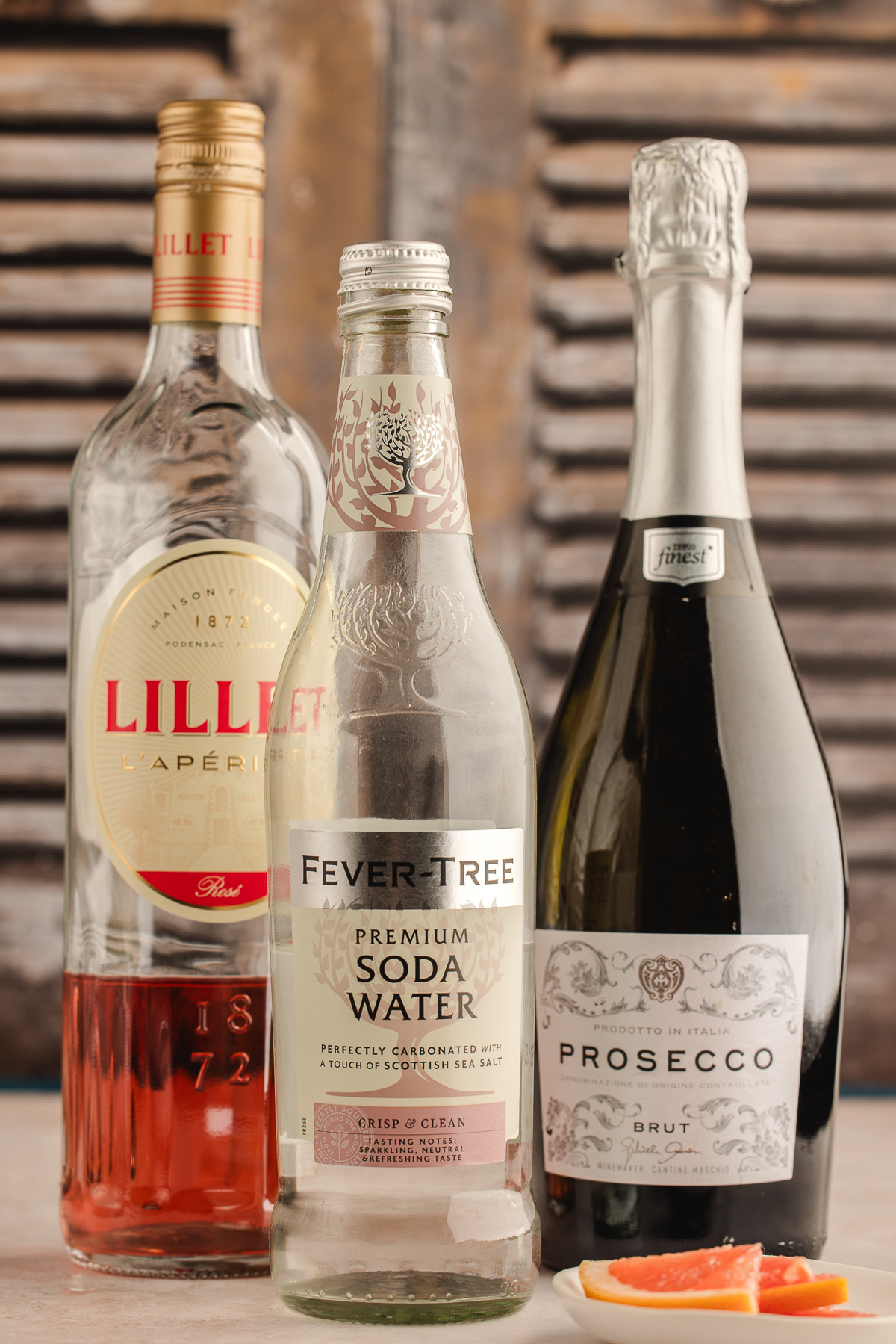 The three ingredients for a Lillet Rosé spritz—Lillet Rosé, club soda, and a bottle of brut prosecco—standing together with fresh grapefruit slices nearby.