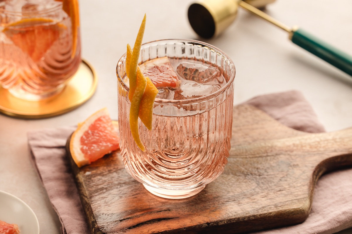 A pale pink Lillet Rosé spritz served over ice in a textured glass, garnished with a long grapefruit twist and set on a wooden board with fresh grapefruit nearby. 