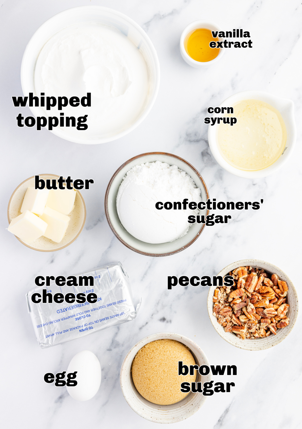 Ingredients for a cream cheese dessert dip arranged on a marble surface, including whipped topping, brown sugar, pecans, confectioners’ sugar, butter, corn syrup, vanilla extract, and an egg.