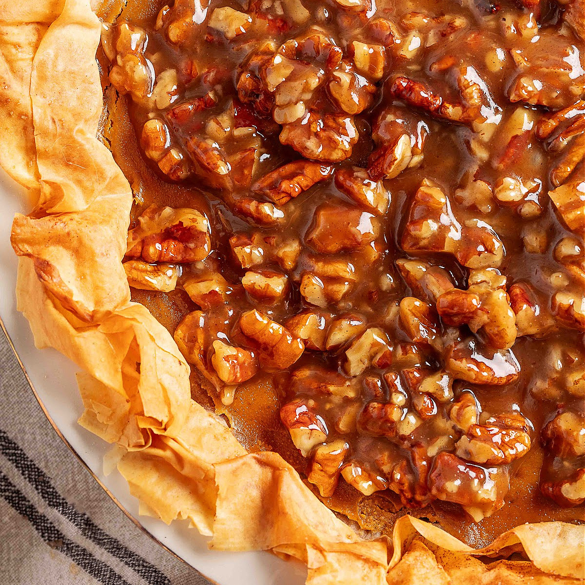 An overhead close-up of pumpkin praline pie showing crisp, ruffled phyllo layers and a glossy brown-sugar pecan praline topping pooled across the surface.