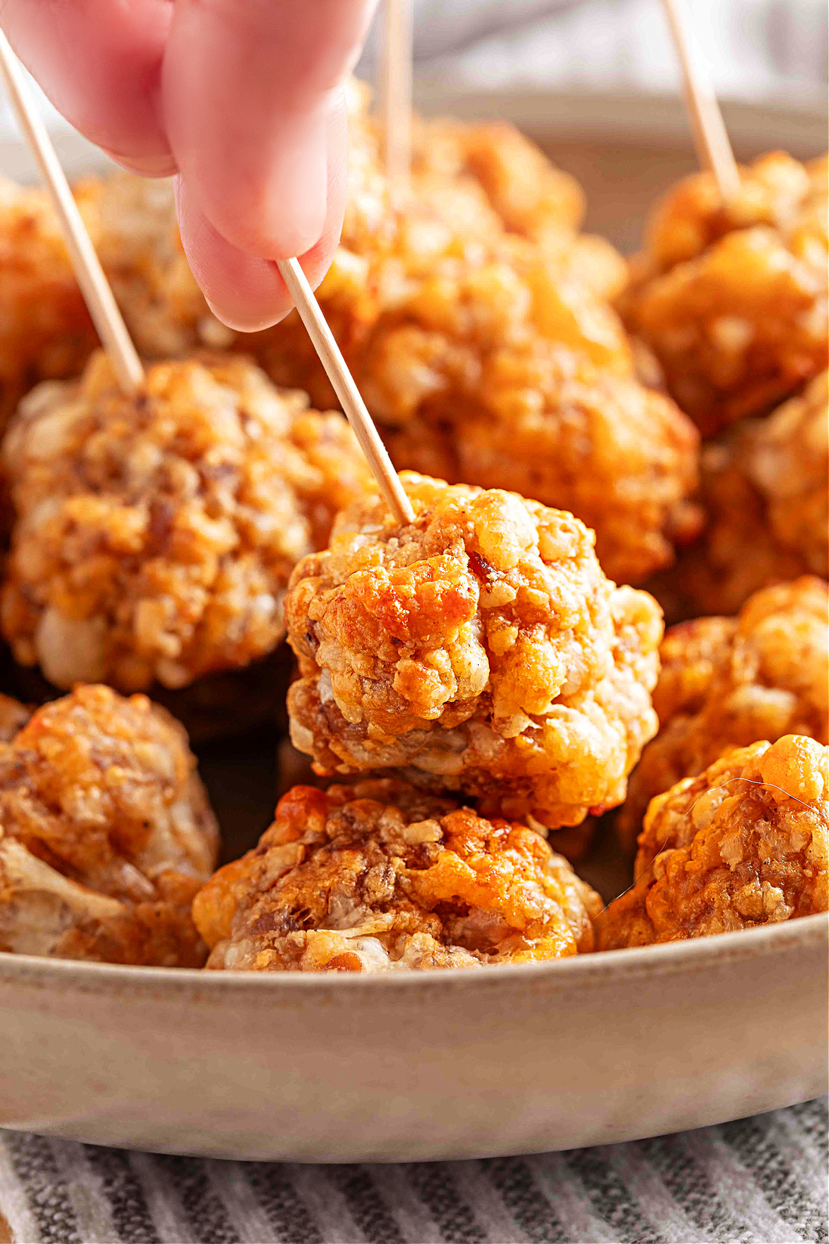 A hand picks up a smoked cheddar sausage ball with a toothpick, showing its crisp, golden surface and tender baked texture, by Noble Pig.