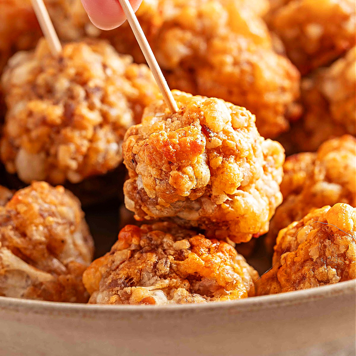 A square, close-up image of sausage balls in a serving bowl, with one lifted on a toothpick to show the texture and color of the smoky, cheesy mixture.