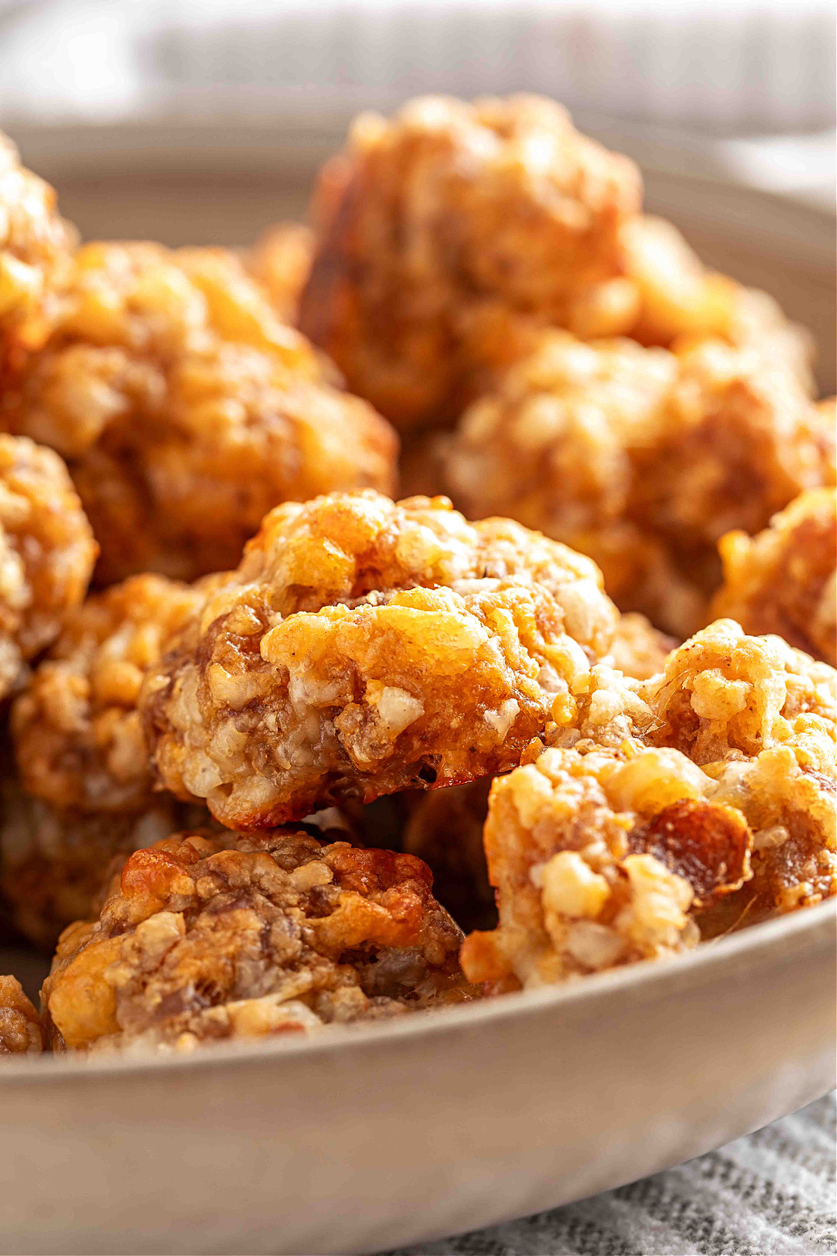 A close-up view of baked sausage balls piled in a shallow bowl, showing their crisp edges, melted smoked cheddar, and golden-brown surface.