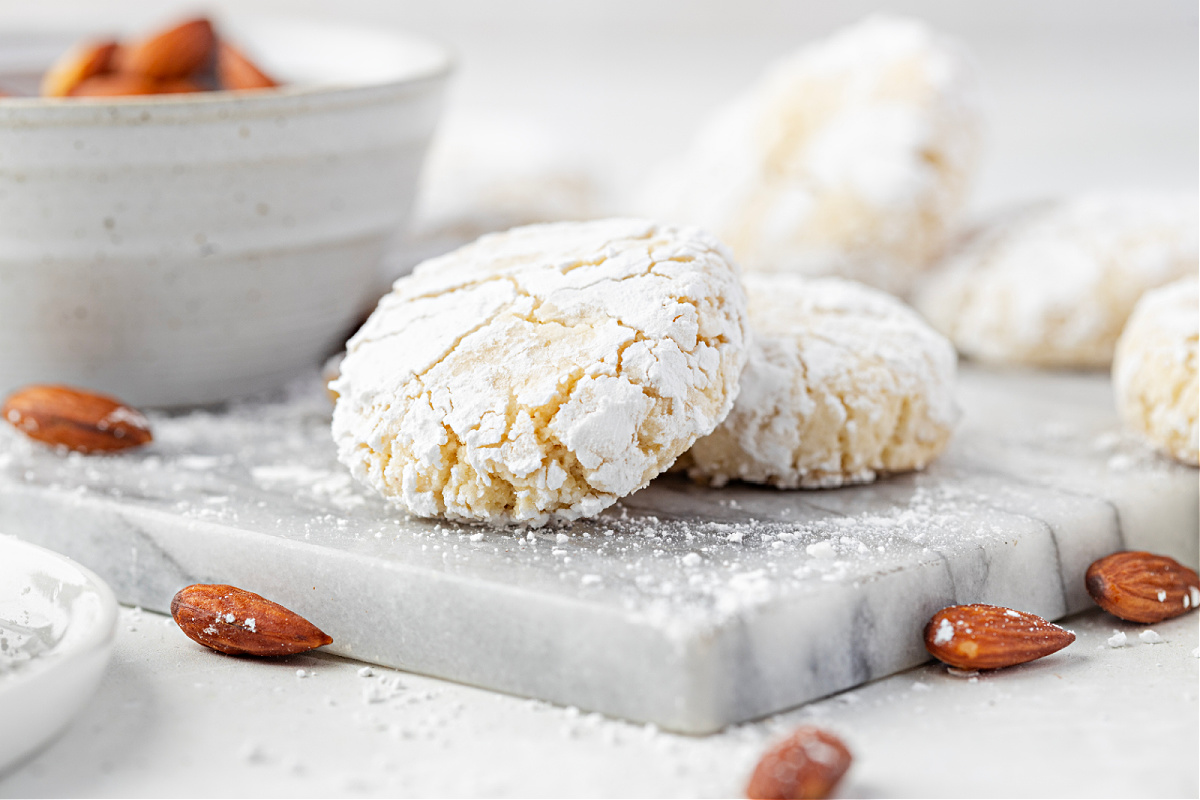Soft amaretti cookies coated in powdered sugar, showing their snowy crackle beside a bowl of almonds.