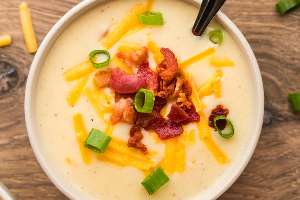 A bowl of creamy baked potato soup topped with crisp bacon, shredded cheddar, and sliced scallions.