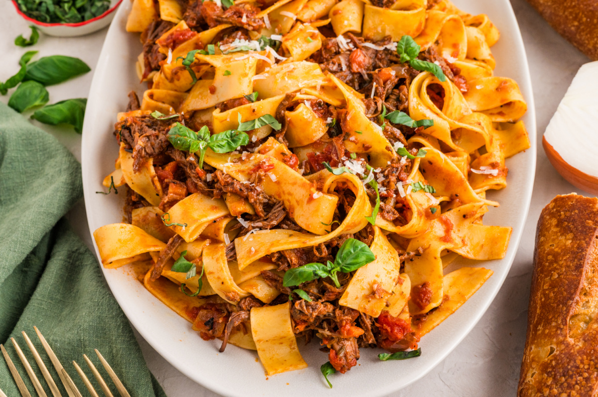 A serving platter of pappardelle tossed with braised beef ragù and fresh basil. A large oval platter filled with tender beef ragù pappardelle, wide noodles twisted with shredded braised beef, tomato-rich sauce, fresh basil, and a sprinkling of parmesan.