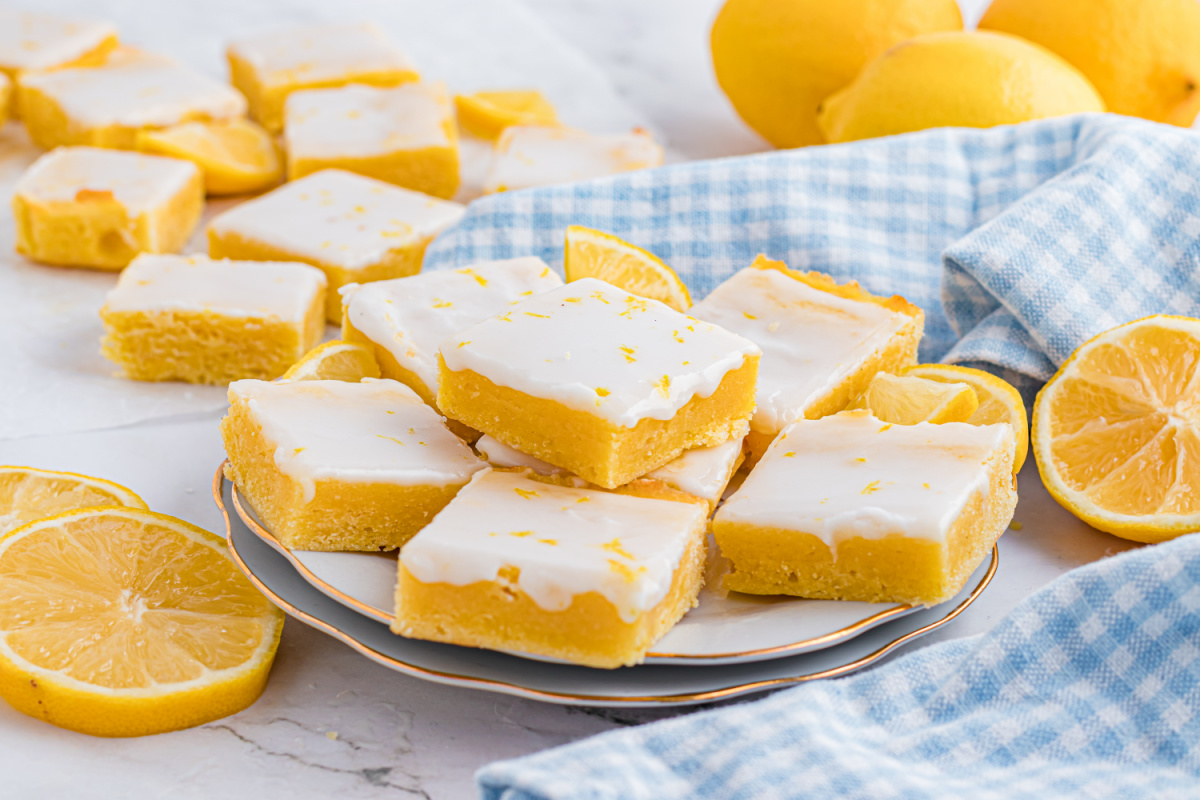 Stacked lemon dessert bars with a thick, buttery crumb and a glossy lemon glaze on top. Close-up of glazed citrus dessert bars stacked on a plate, showing a dense lemon crumb and smooth white glaze.