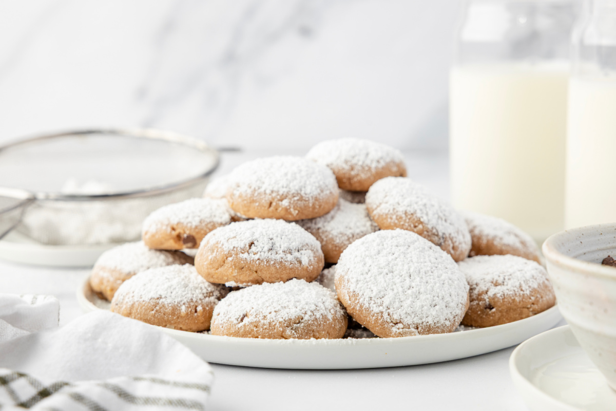 Fresh Danish wedding cookies on a serving plate. A plate of Danish wedding cookies next to a sugar sifter and bottles of milk on a bright countertop.