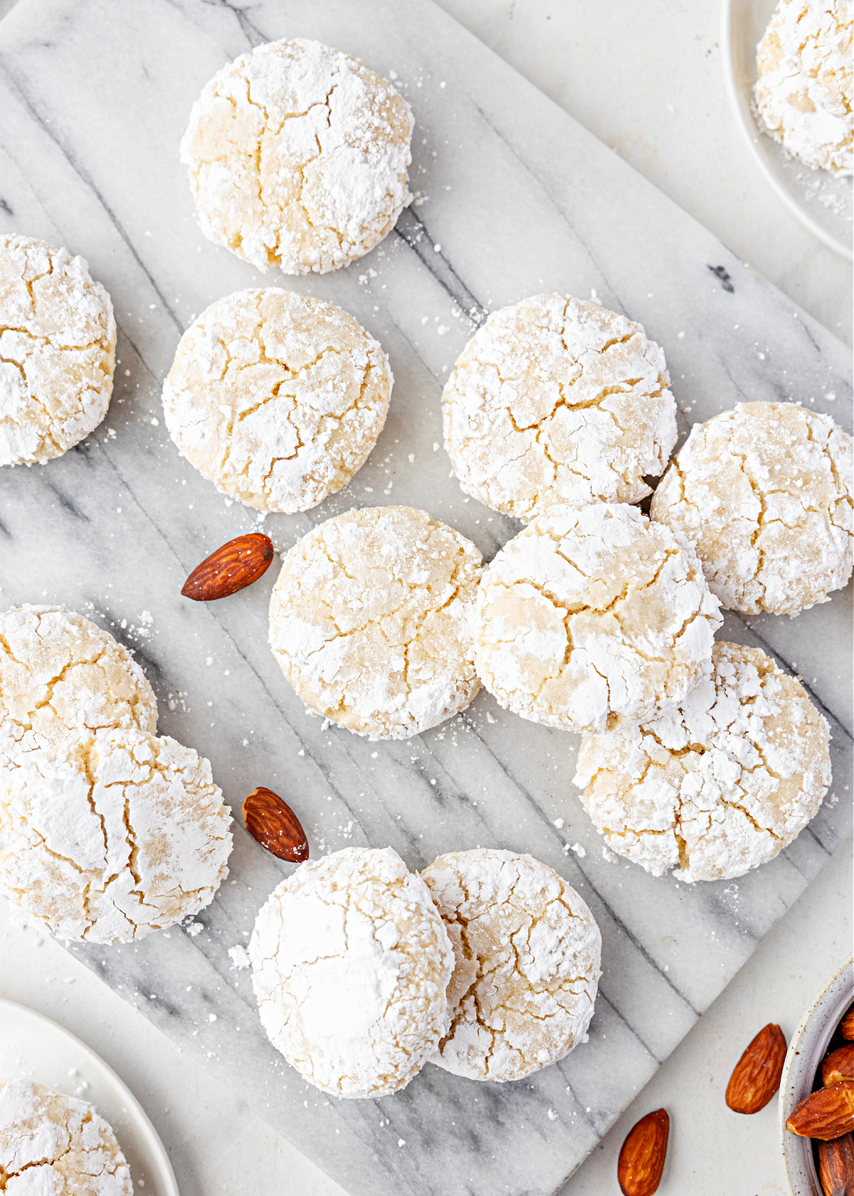 Snowy, crackled almond cookies arranged on a marble board with scattered almonds.