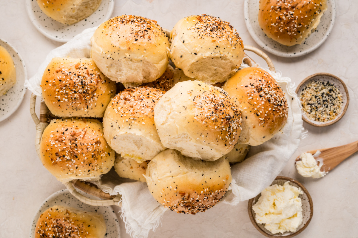 Basket of warm savory rolls with a seasoned crust, served with cream cheese and everything seasoning on the side. A basket filled with freshly baked rolls topped with sesame seeds, poppy seeds, garlic, and onion, surrounded by small plates of cream cheese and extra seasoning.