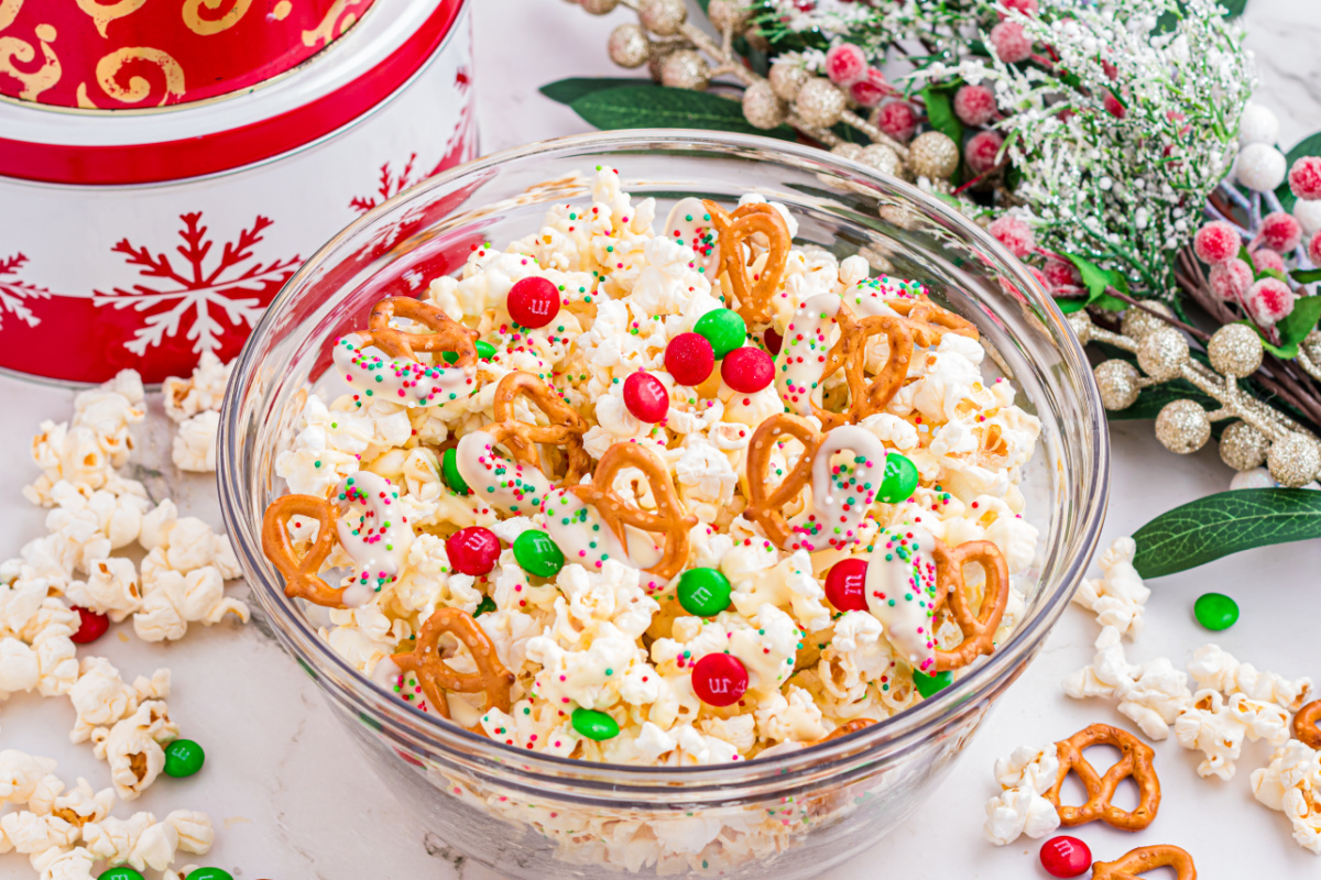 Holiday popcorn mix with white chocolate–dipped pretzels, red and green candy-coated chocolates, and sprinkles in a glass bowl.