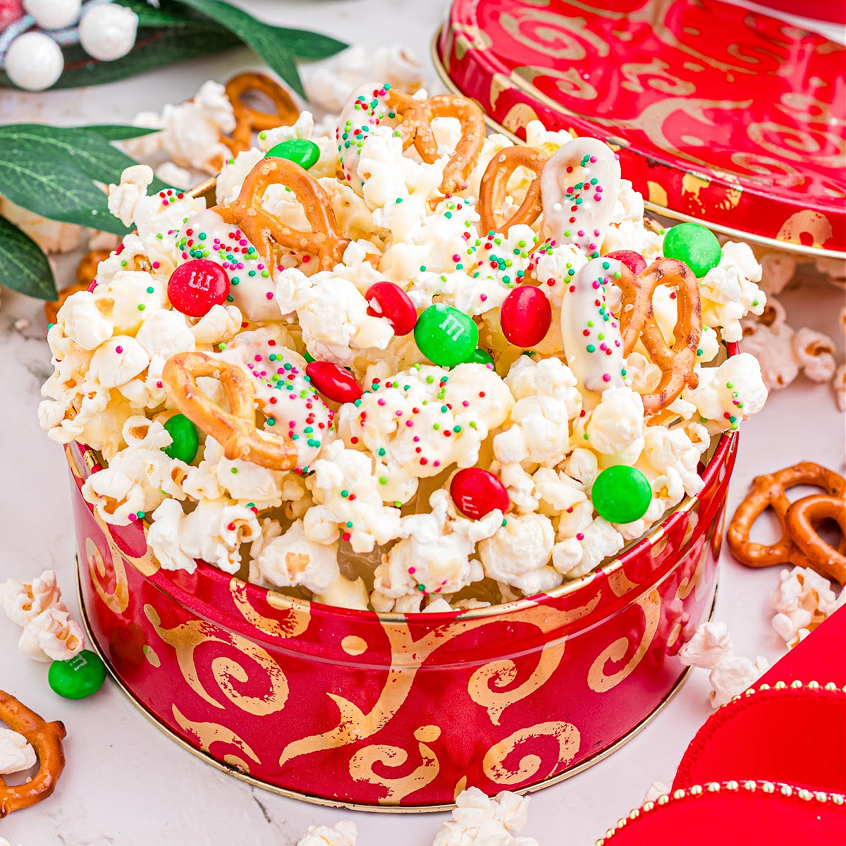 Christmas popcorn mix piled into a red holiday tin with white chocolate–dipped pretzels, candy-coated chocolates, and festive sprinkles.