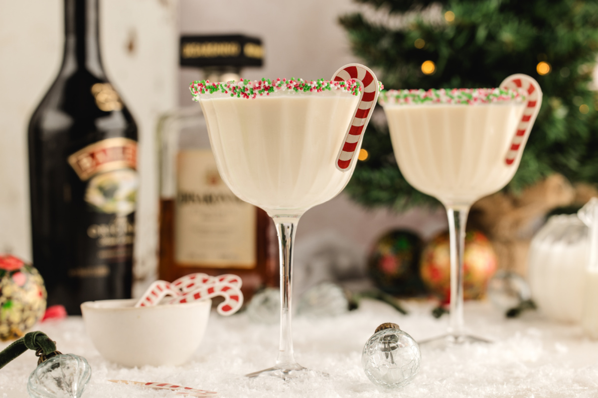 Two sprinkle-rimmed holiday martinis with candy cane stirrers in a festive winter scene. Two creamy holiday martinis with sprinkle rims and candy cane stirrers set on a snowy tabletop, with liqueur bottles and a Christmas tree softly blurred in the background.