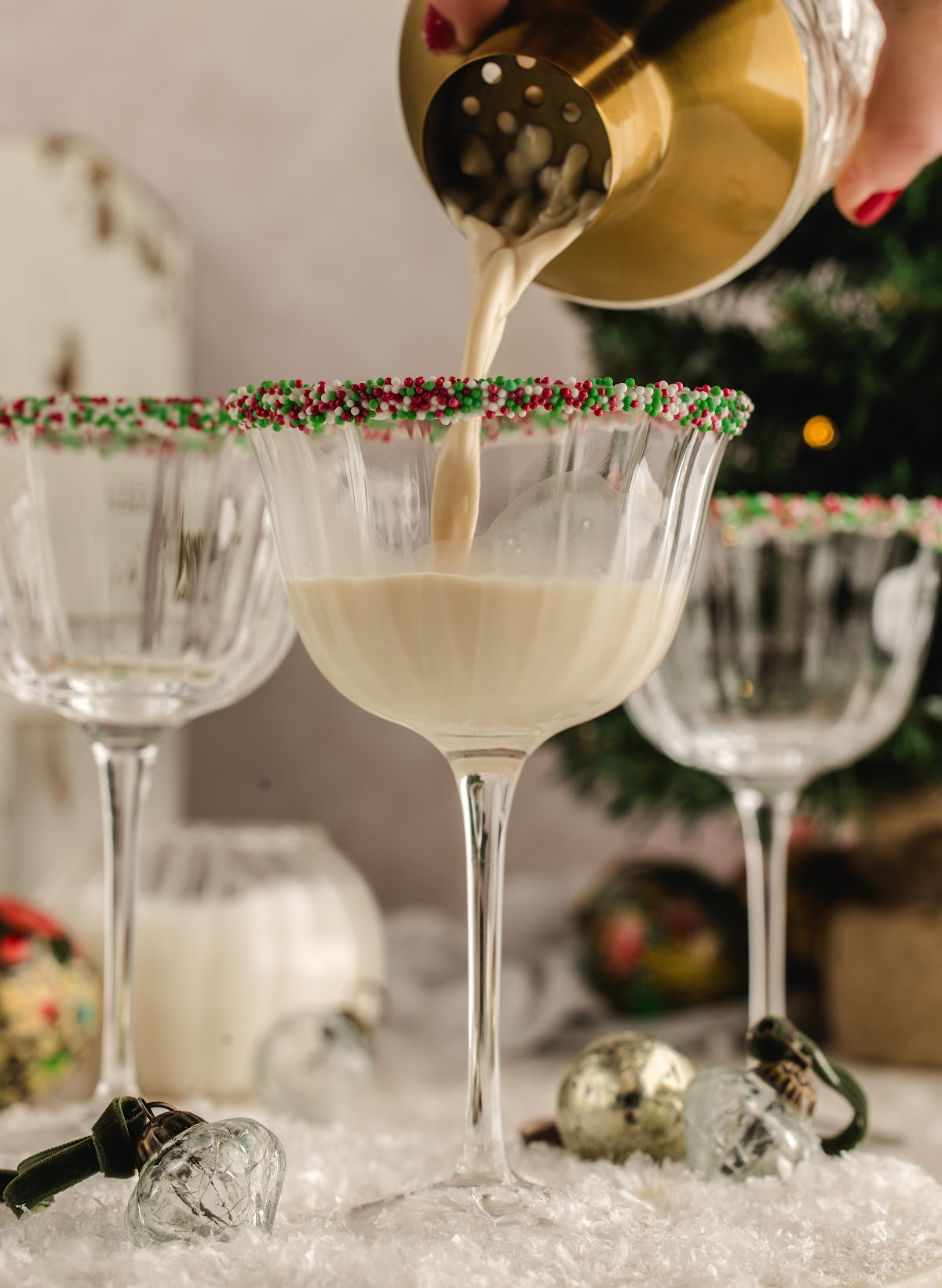A festive cream cocktail poured into a sprinkle-rimmed coupe in a holiday scene. A creamy holiday cocktail being strained from a gold shaker into a sprinkle-rimmed coupe glass, surrounded by ornaments and snowy decorations.