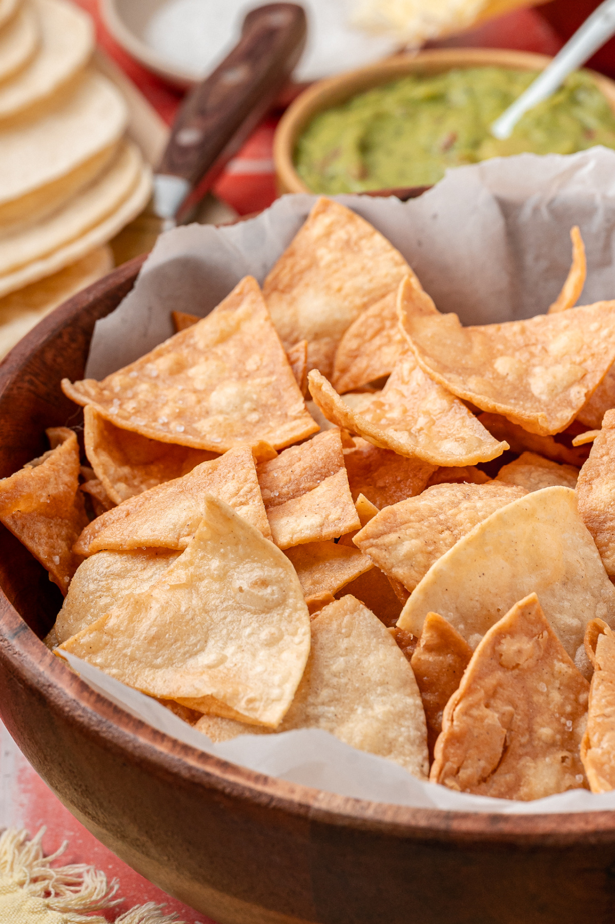 Close-up view of freshly fried corn tortilla chips resting in a wooden bowl. A close-up of golden, blistered corn tortilla chips piled in a wooden bowl, showing crisp edges and light salt clinging to the surface.