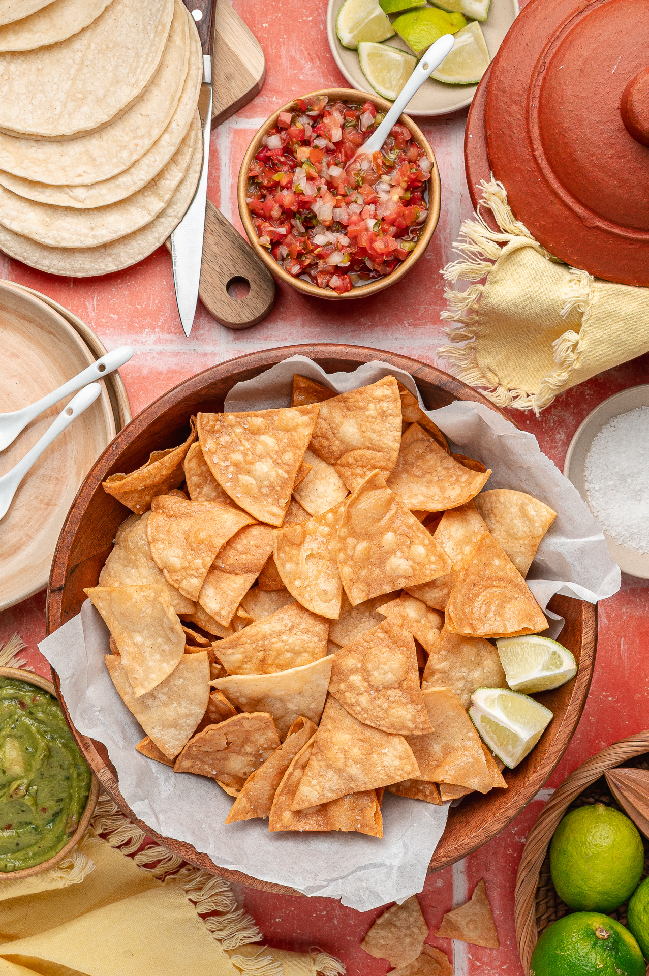 A bowl of freshly fried tortilla chips set out with salsa, guacamole, and lime wedges for serving. A large wooden bowl of easy fried tortilla chips piled high with blistered, golden wedges, surrounded by fresh salsa, guacamole, and lime.