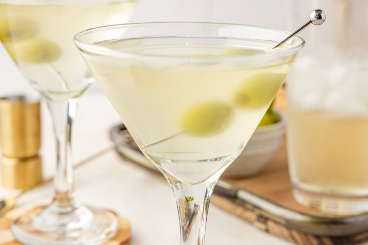 Martinis made with olive brine and green olives, served ice-cold with a shaker and bar tools nearby. Two chilled martinis with green olives resting in pale, briny cocktails, set beside a shaker and a small bowl of olives.