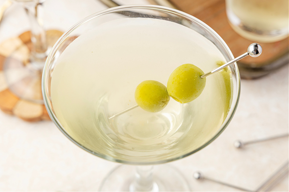 Vodka martini made with olive brine and green olives, served ice-cold in a stemmed glass. A chilled martini glass filled with a pale, briny cocktail and green olives, set against a shaker and bowls of olives in the background.