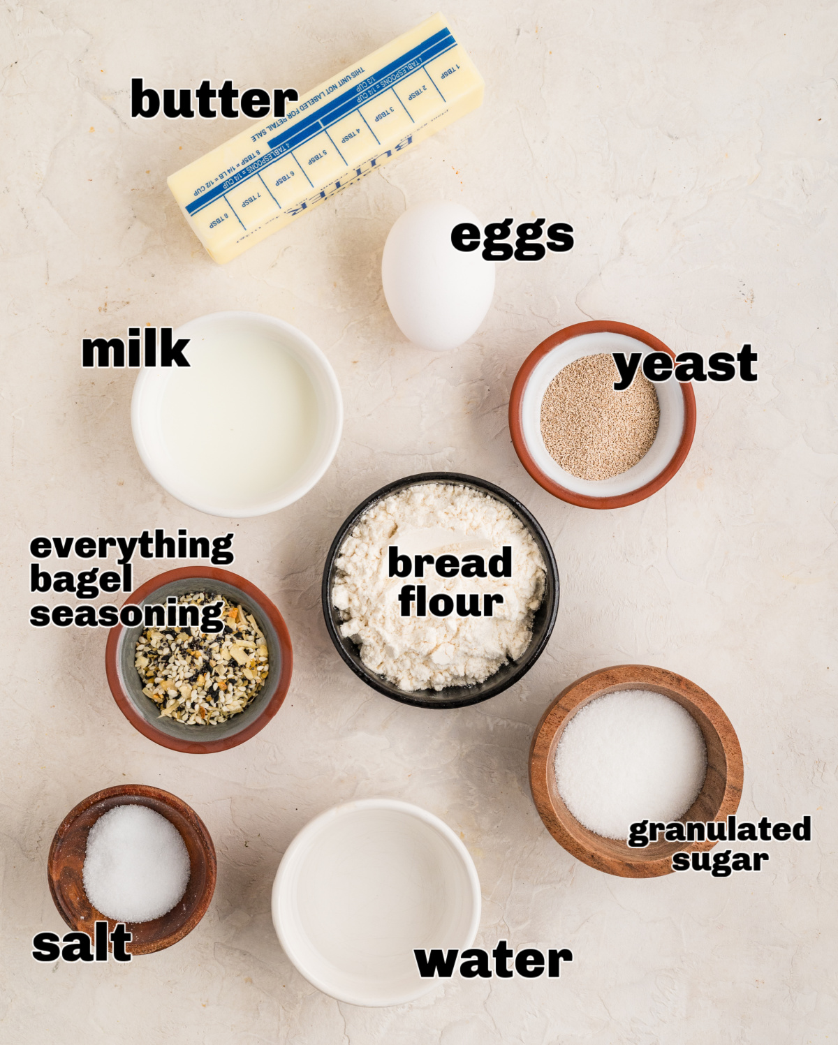 Ingredients laid out for making seasoned yeast rolls, photographed overhead with flour, yeast, dairy, eggs, and everything seasoning. Measured ingredients for savory yeast rolls arranged on a light surface, including bread flour, yeast, eggs, milk, butter, sugar, salt, and everything bagel seasoning.