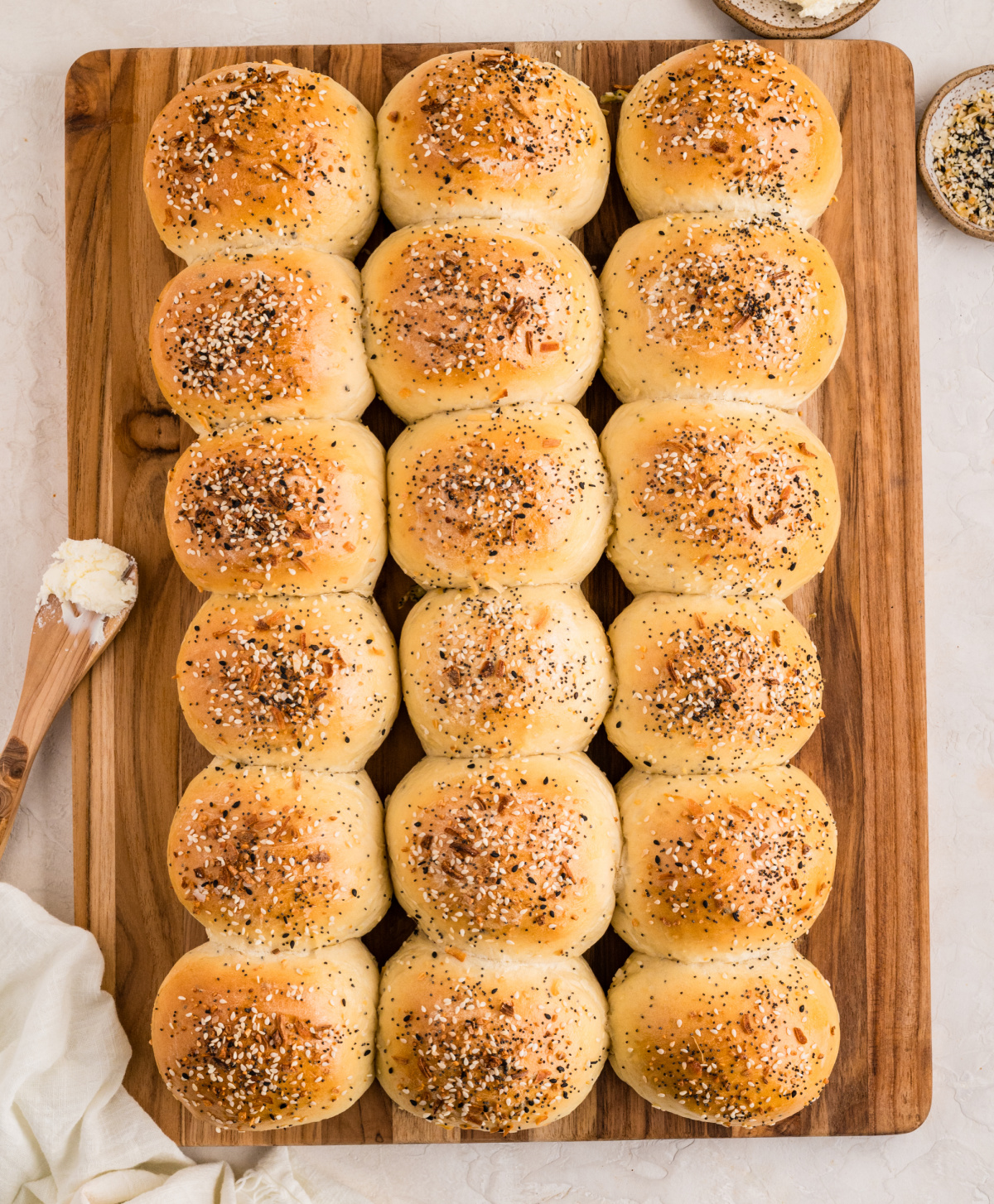 Freshly baked savory rolls with a seasoned crust, photographed overhead on a wooden board. A full tray of golden baked rolls arranged in neat rows on a wooden board, topped with sesame seeds, poppy seeds, garlic, and onion seasoning.