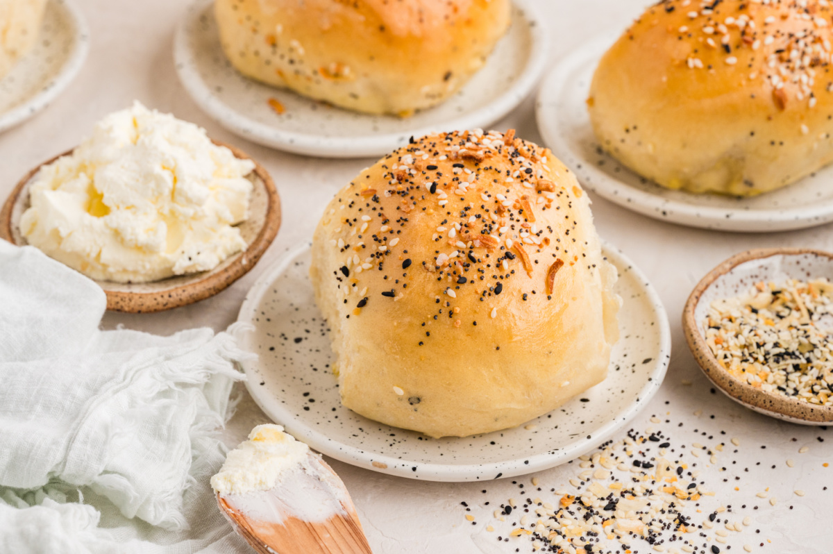Savory seeded rolls served warm with cream cheese and scattered everything seasoning on a light surface. Soft baked rolls topped with sesame seeds, poppy seeds, garlic, and onion, served on small plates with cream cheese and extra seasoning nearby.