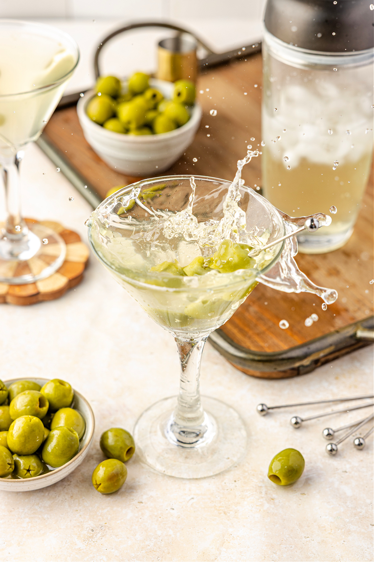 Shaken martini splashing with olive brine and green olives, surrounded by cocktail tools and ice. A chilled martini splashing dramatically in the glass as green olives tumble through the pale, briny cocktail on a bar tray.