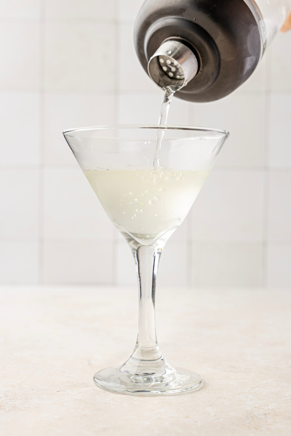 Straining a shaken olive brine martini into a cold glass, showing the cloudy, saline finish from shaking. A pale, briny martini being strained from a shaker into a chilled stemmed glass.