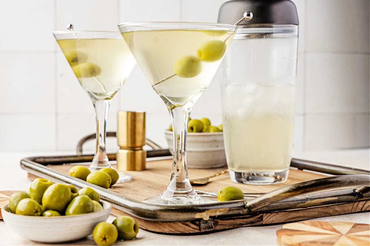 Vodka martinis made with olive brine, served ice-cold with green olives and bar tools nearby. Two briny martinis garnished with green olives on a tray beside a cocktail shaker and bowls of olives.