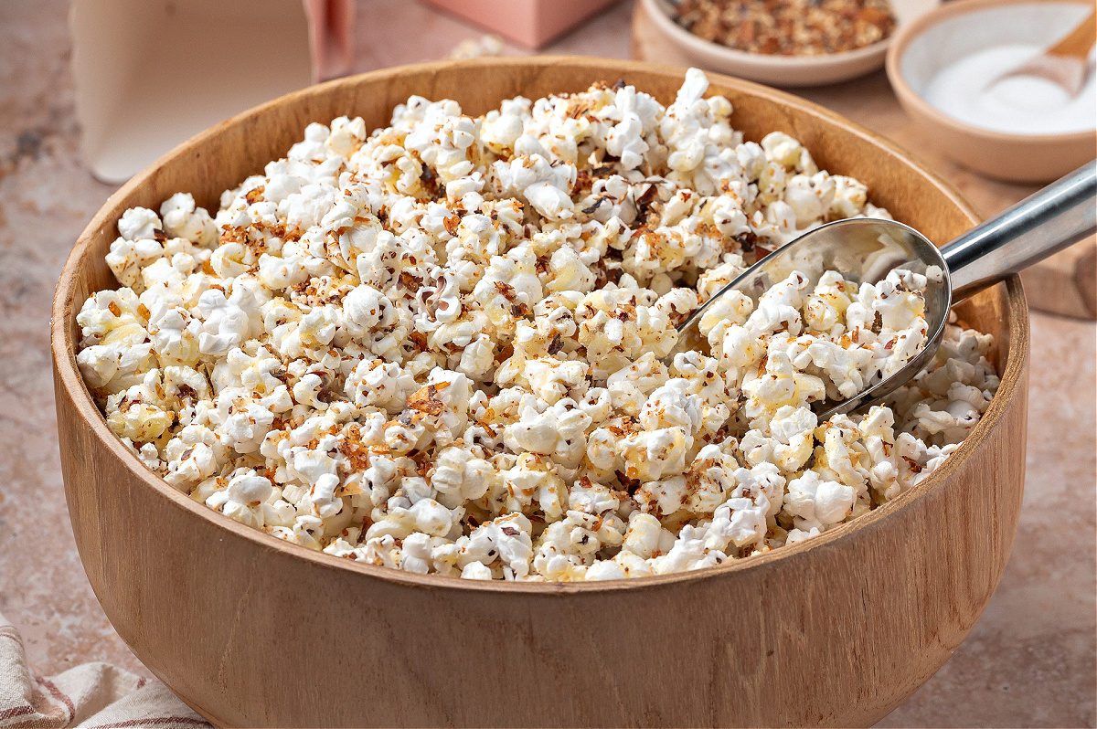 A bowl of seasoned popcorn with a metal scoop. A wooden bowl filled with popcorn coated in a sweet and spicy seasoning blend, with a metal scoop resting inside.