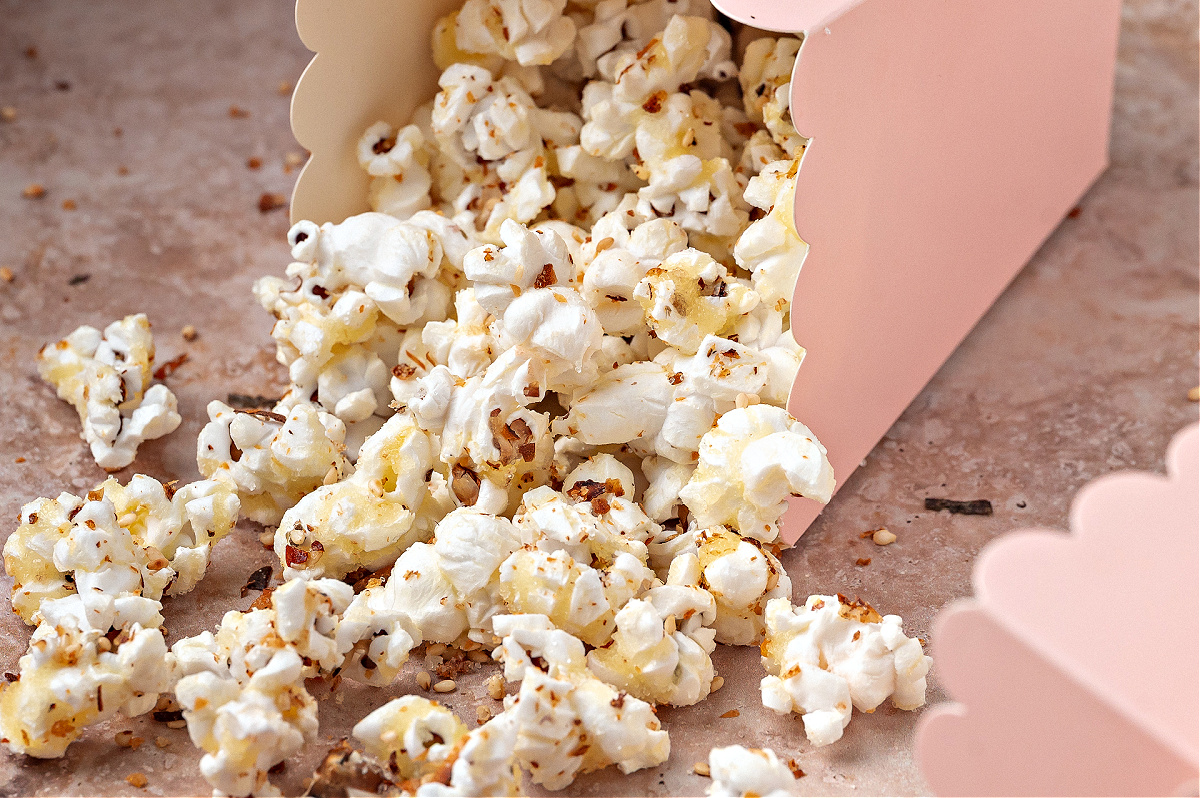 Furikake popcorn tumbling out of a pink treat box onto the surface. Sweet and spicy furikake popcorn spilling from a pink scalloped treat box, showing the toasted seasoning clinging to each kernel.