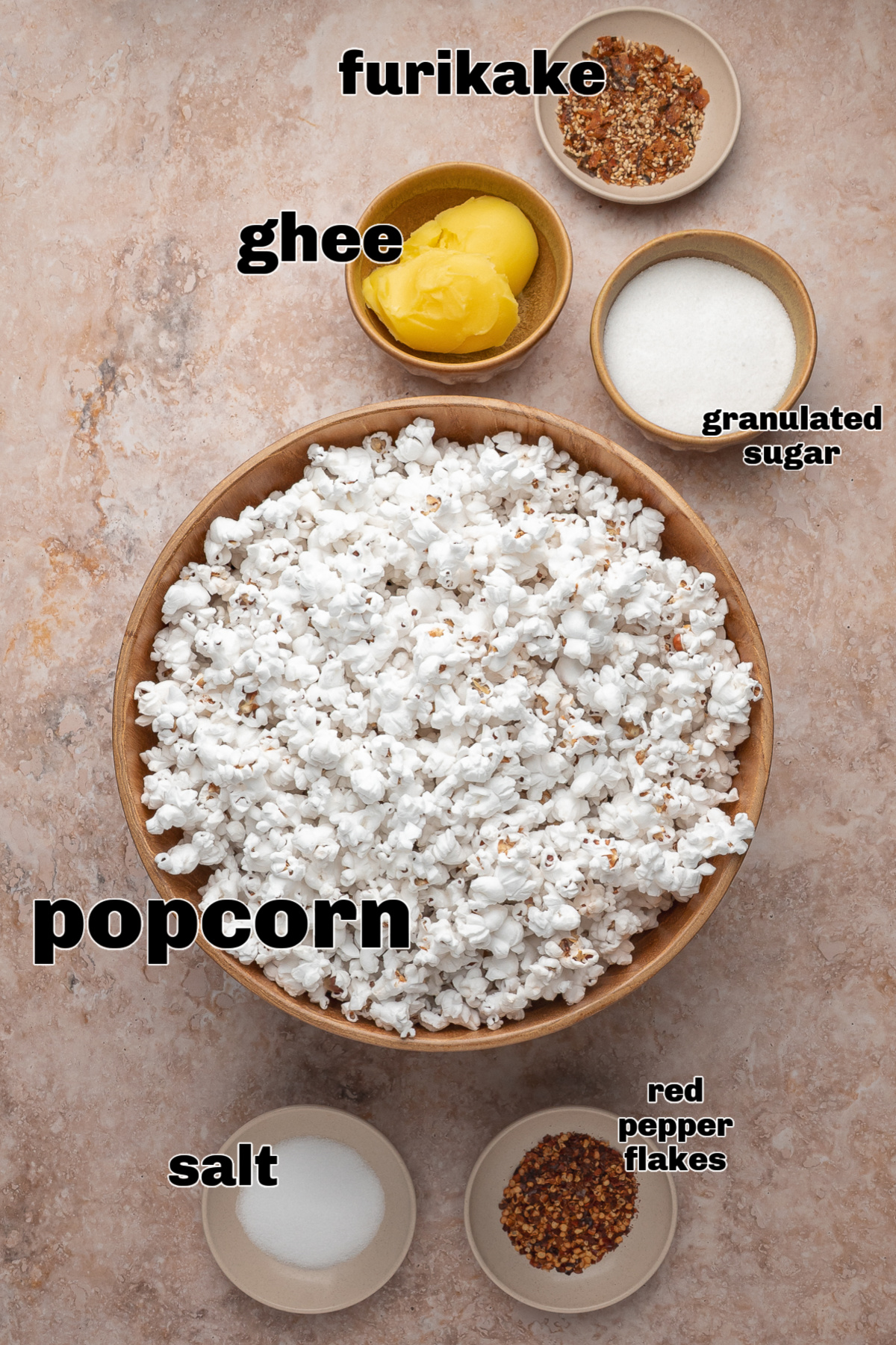 Ingredients for the seasoned popcorn arranged in bowls around a large bowl of popped corn. A flat lay of the popcorn ingredients arranged around a large wooden bowl, including ghee, sugar, salt, red pepper flakes, and furikake seasoning.
