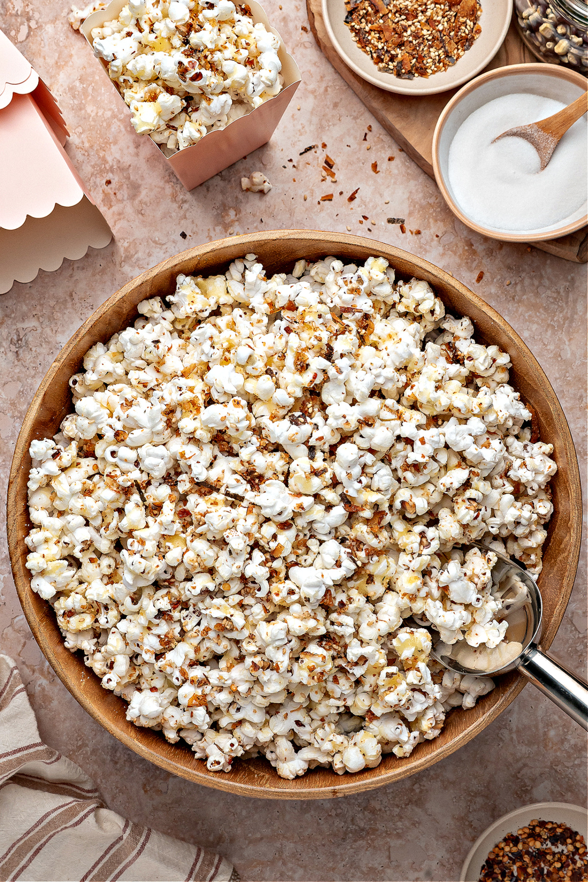 A wooden bowl filled with seasoned popcorn surrounded by small dishes of ingredients. A large wooden bowl of sweet and spicy furikake popcorn sits on a stone surface, with scattered seasoning, small serving containers, and ingredients visible around it.
