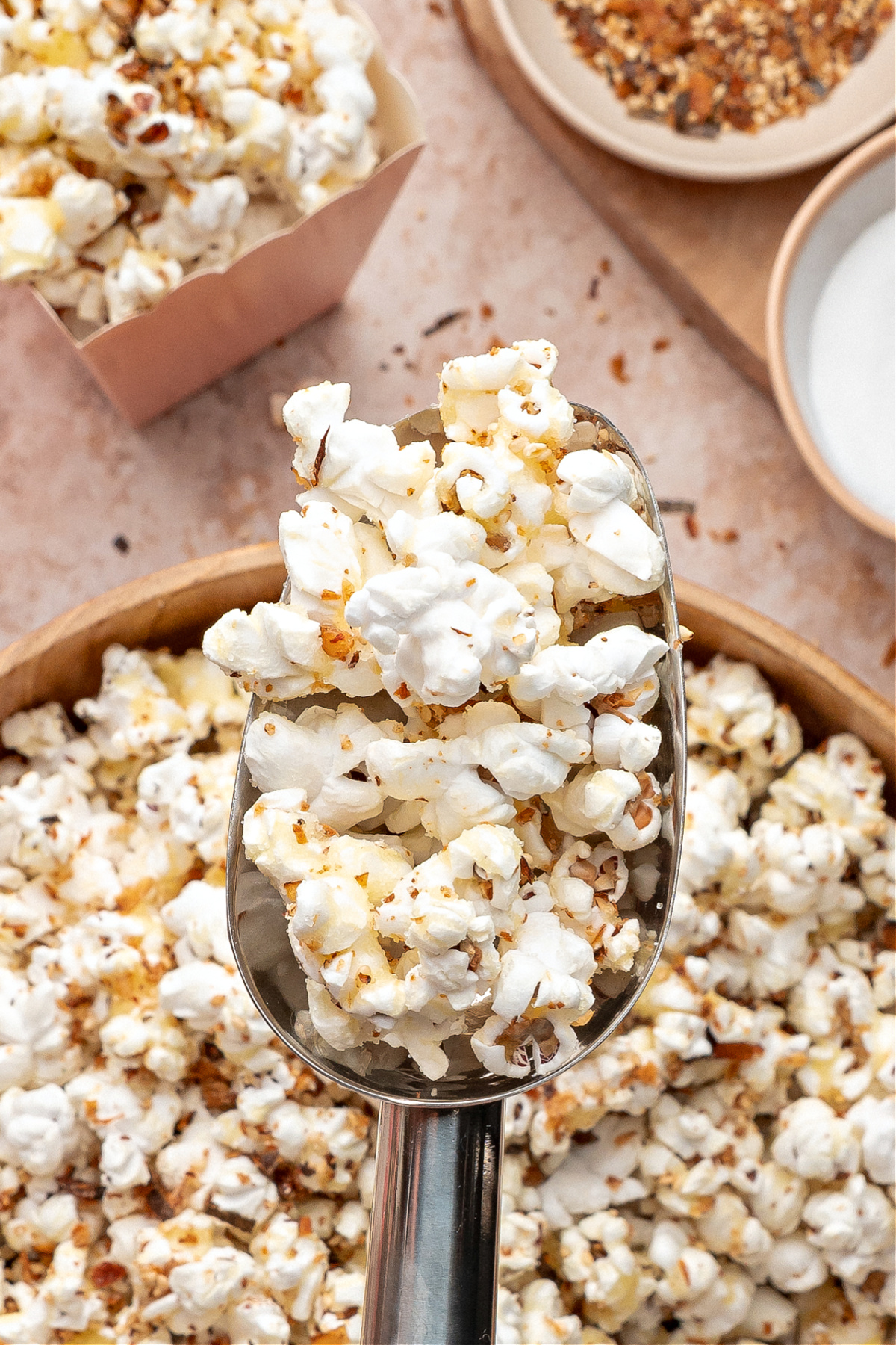 Scoop of flavored popcorn held above a bowl with seasoning dishes nearby. A metal scoop lifting a serving of seasoned popcorn above a wooden bowl, with toppings and ingredients in the background.
