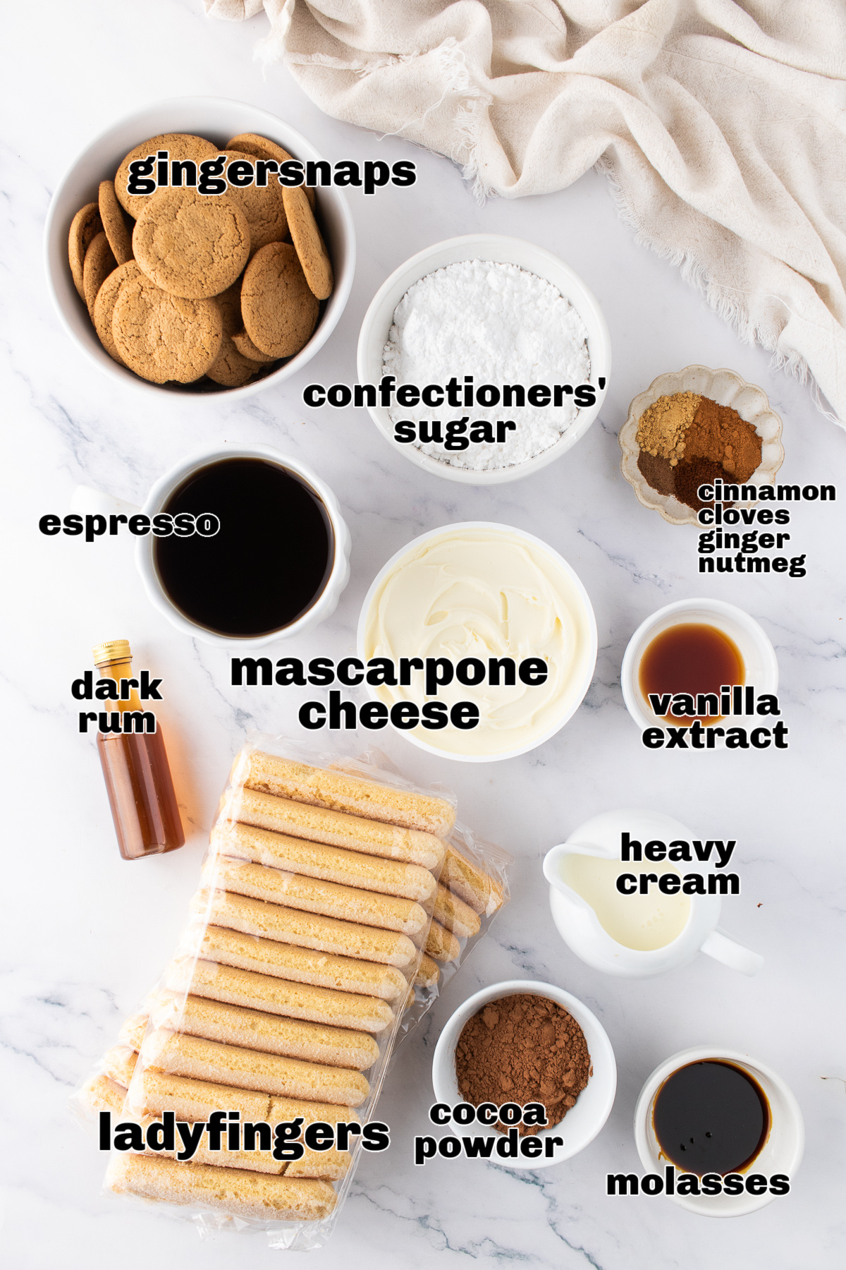 Ingredients arranged for making a spiced gingerbread tiramisu. A top-down view of the gingerbread tiramisu ingredients laid out on a marble surface, including gingersnaps, ladyfingers, mascarpone, espresso, spices, molasses, and confectioners’ sugar.