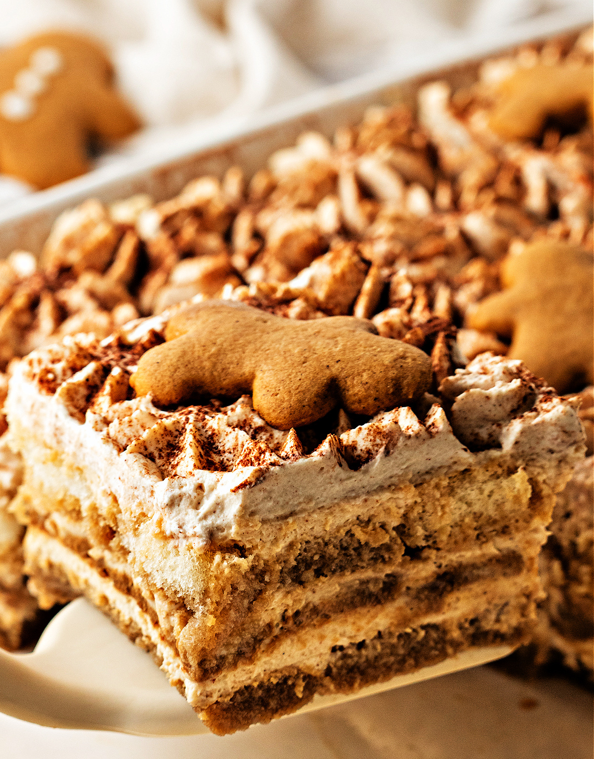 Slice of gingerbread tiramisu with layered espresso-soaked cookies and spiced mascarpone. A close slice of gingerbread tiramisu lifted from the pan, showing soft espresso-soaked layers, spiced mascarpone cream, and a small gingerbread cookie resting on top.