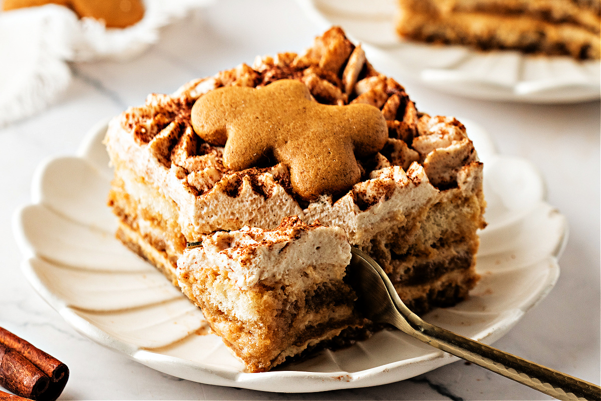 A creamy layered slice of gingerbread tiramisu with a fork taking a bite. A slice of gingerbread tiramisu with a gingerbread cookie on top, showing the creamy spiced layers and a fork taking the first bite on a scalloped plate.