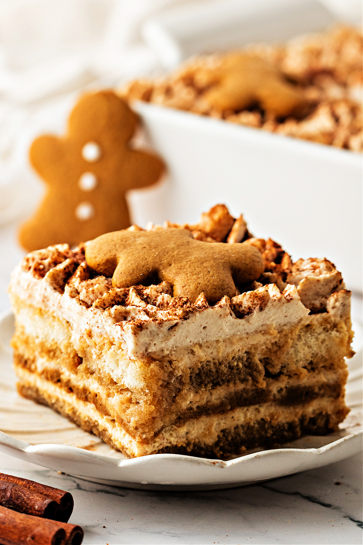 A slice of gingerbread tiramisu topped with a gingerbread cookie on a white plate. A close slice of gingerbread tiramisu on a scalloped plate, layered with espresso-soaked ladyfingers and spiced mascarpone, topped with a small gingerbread cookie and dusted with cocoa.