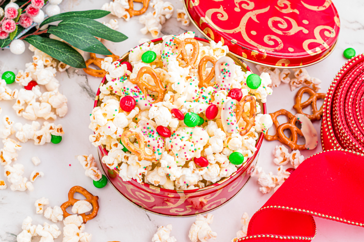 Festive popcorn mix with white chocolate–dipped pretzels, red and green candies, and sprinkles in a holiday tin.