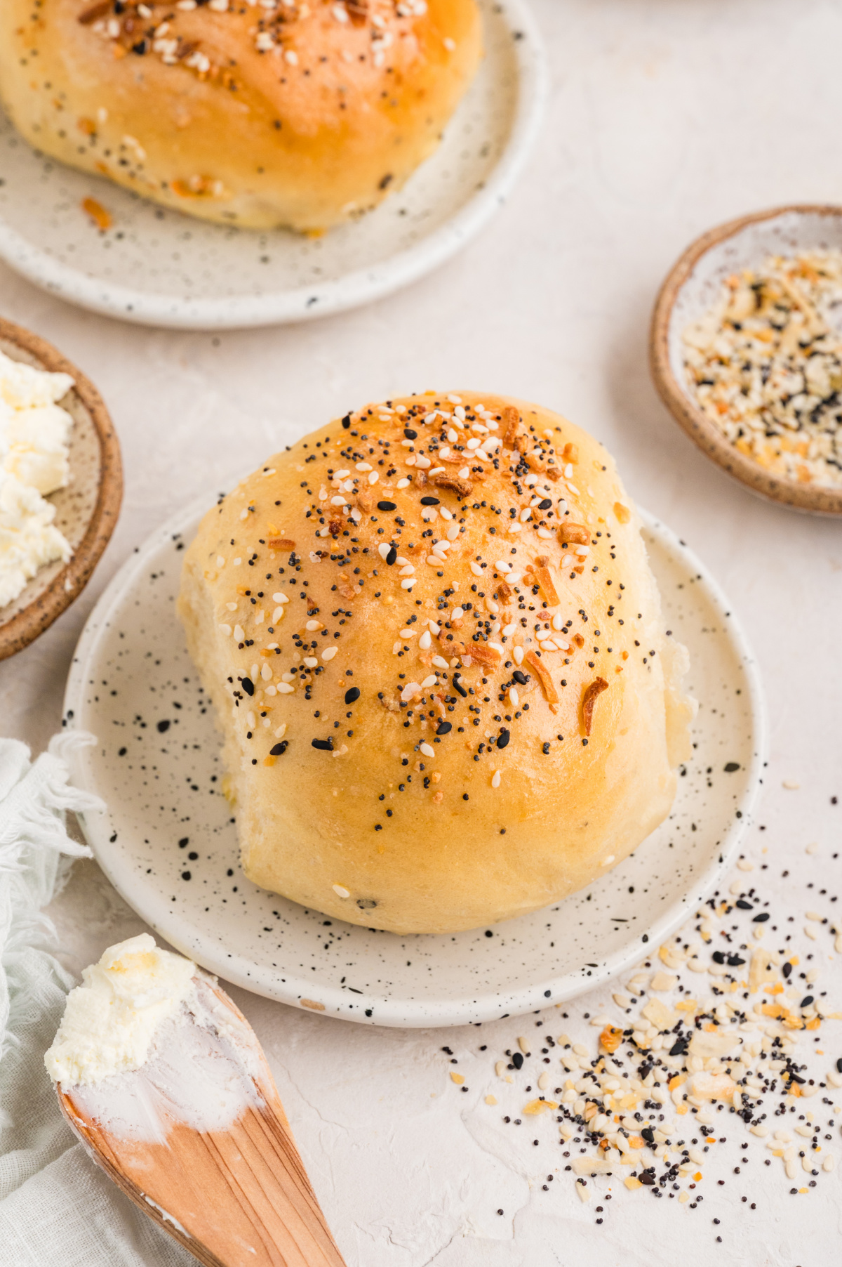 Close-up of a warm savory roll with everything-style seasoning, served simply with cream cheese on the side. A single golden baked roll topped with sesame seeds, poppy seeds, garlic, and onion, resting on a small plate with cream cheese and loose seasoning nearby.