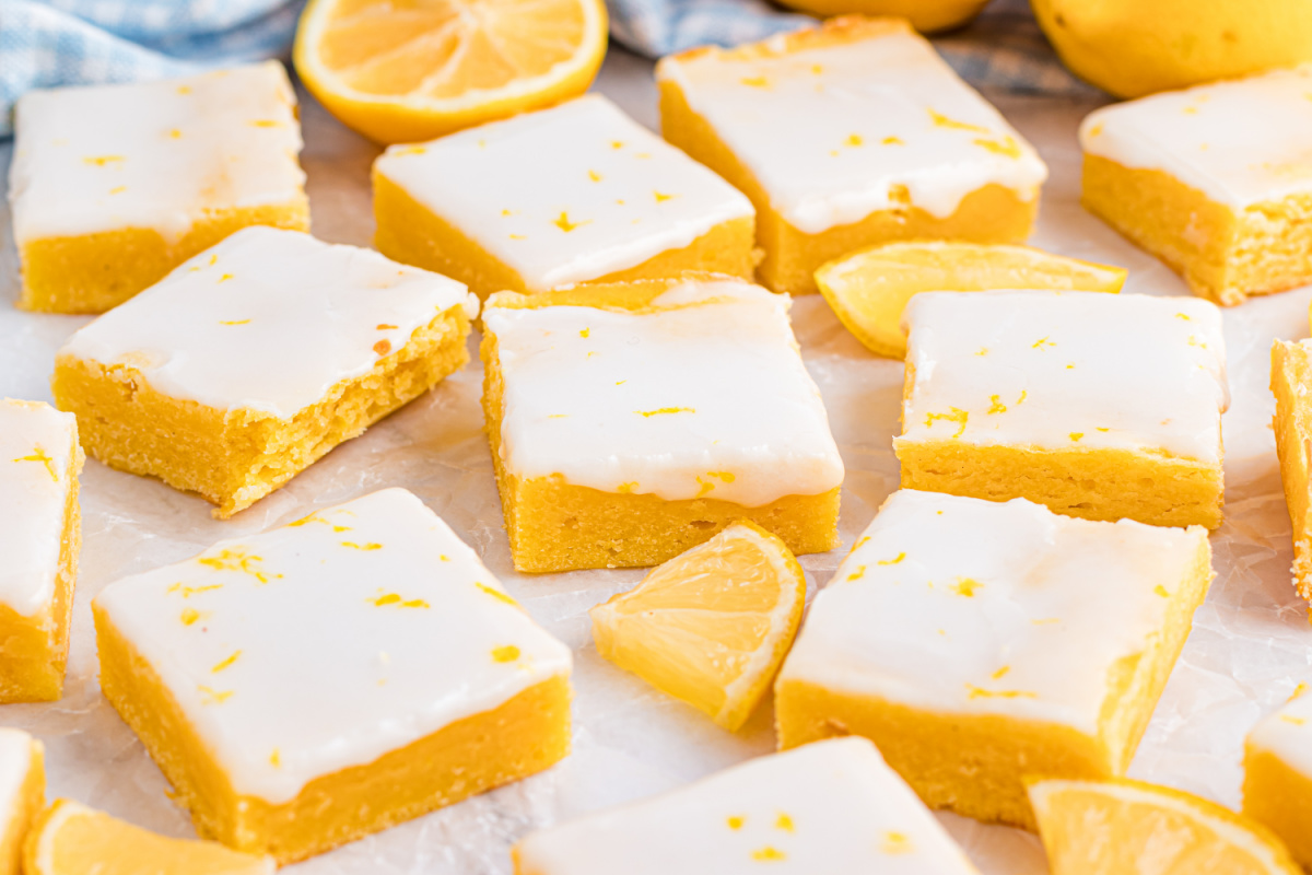 Finished lemon blondies styled with whole lemons and parchment for a clean, bright presentation Lemon blondies arranged in a loose grid on parchment with fresh lemons nearby, highlighting the pale glaze and bright citrus color.
