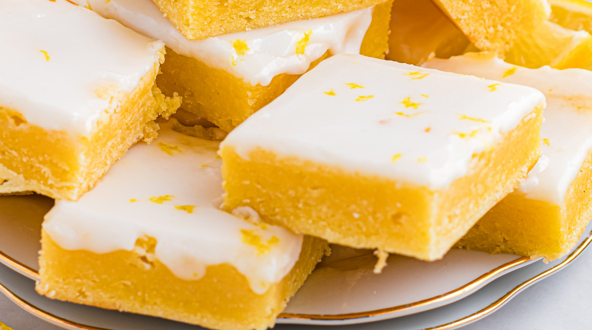 Plated lemon dessert squares with a thin glaze, fresh lemon wedges, and a casual kitchen setting. Lemon blondies cut into neat squares with a smooth lemon glaze, photographed in bright natural light to highlight the citrus color and texture.