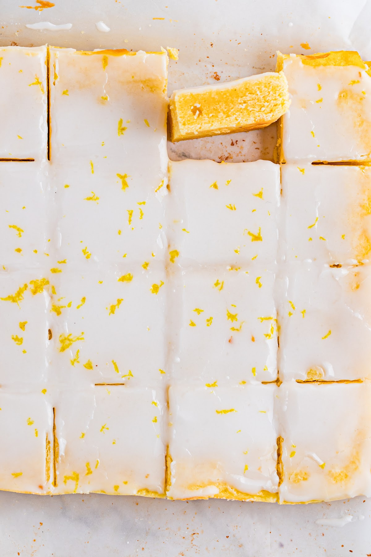 Fully baked blondies sliced in the pan with a set lemon glaze and visible citrus crumb. Overhead view of sliced lemon blondies in the pan, showing a smooth lemon glaze, clean cuts, and a dense yellow crumb.