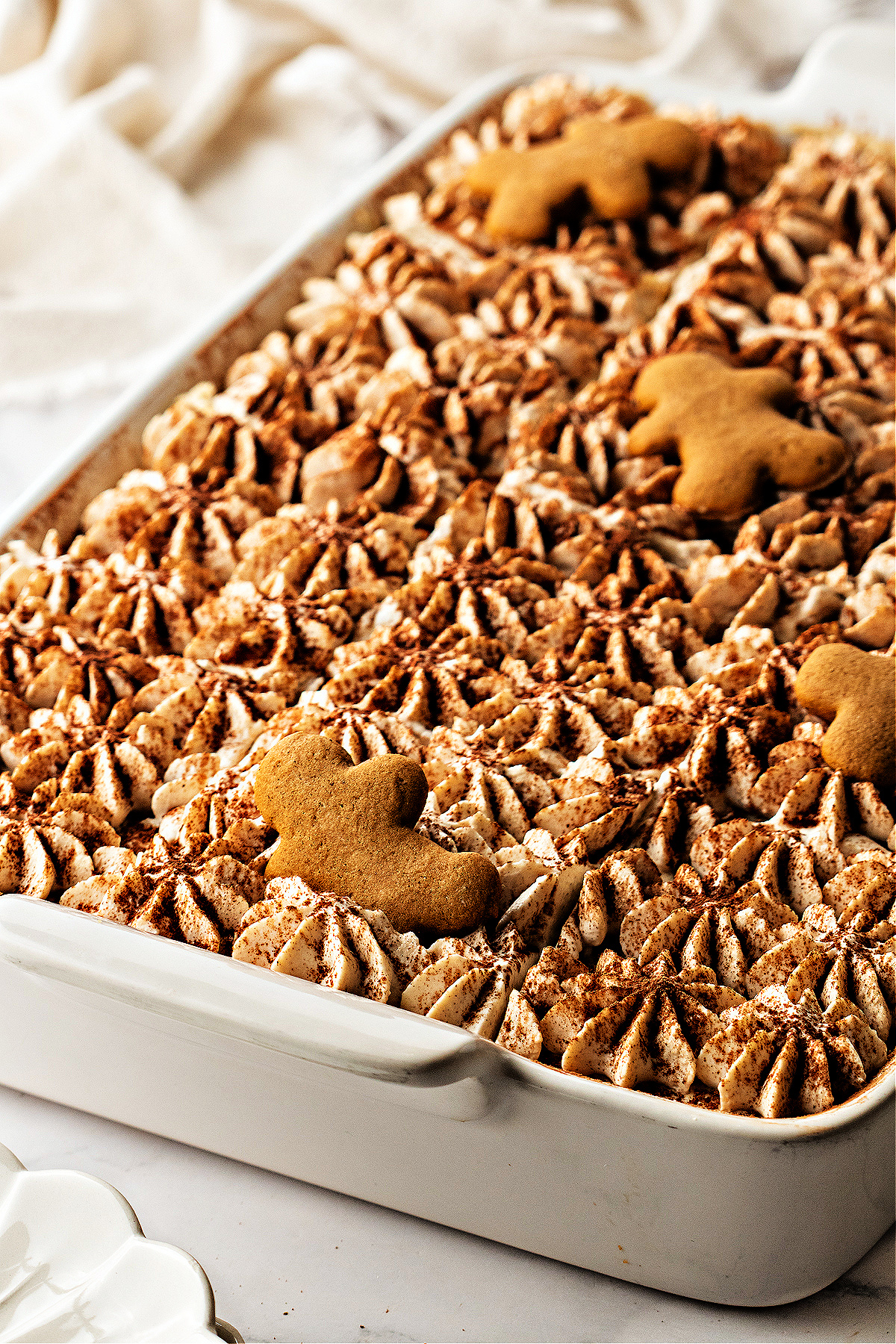 A pan of holiday tiramisu decorated with cocoa and gingerbread cookies. A full pan of spiced holiday tiramisu topped with piped cream, cocoa dusting, and gingerbread men resting across the surface.