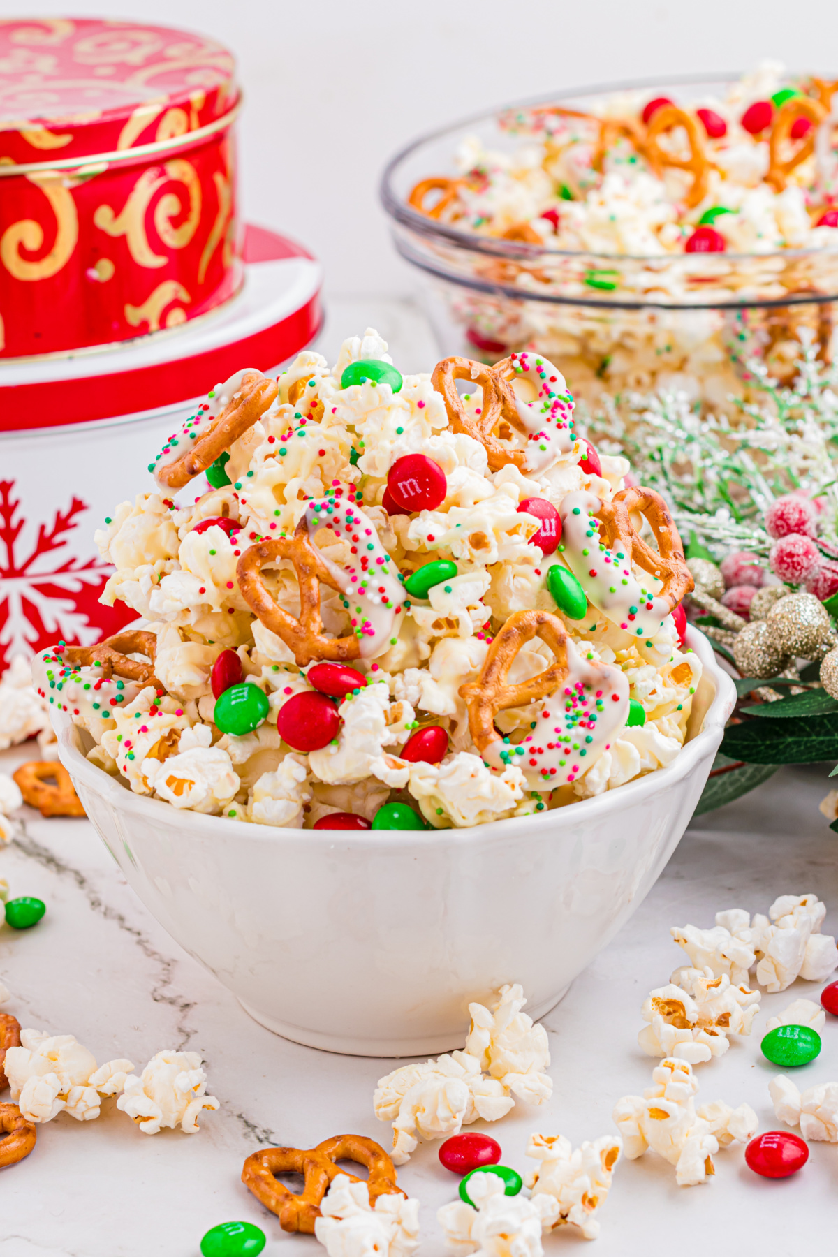 Christmas popcorn mix piled into a white bowl with white chocolate–dipped pretzels, red and green candies, and holiday sprinkles.