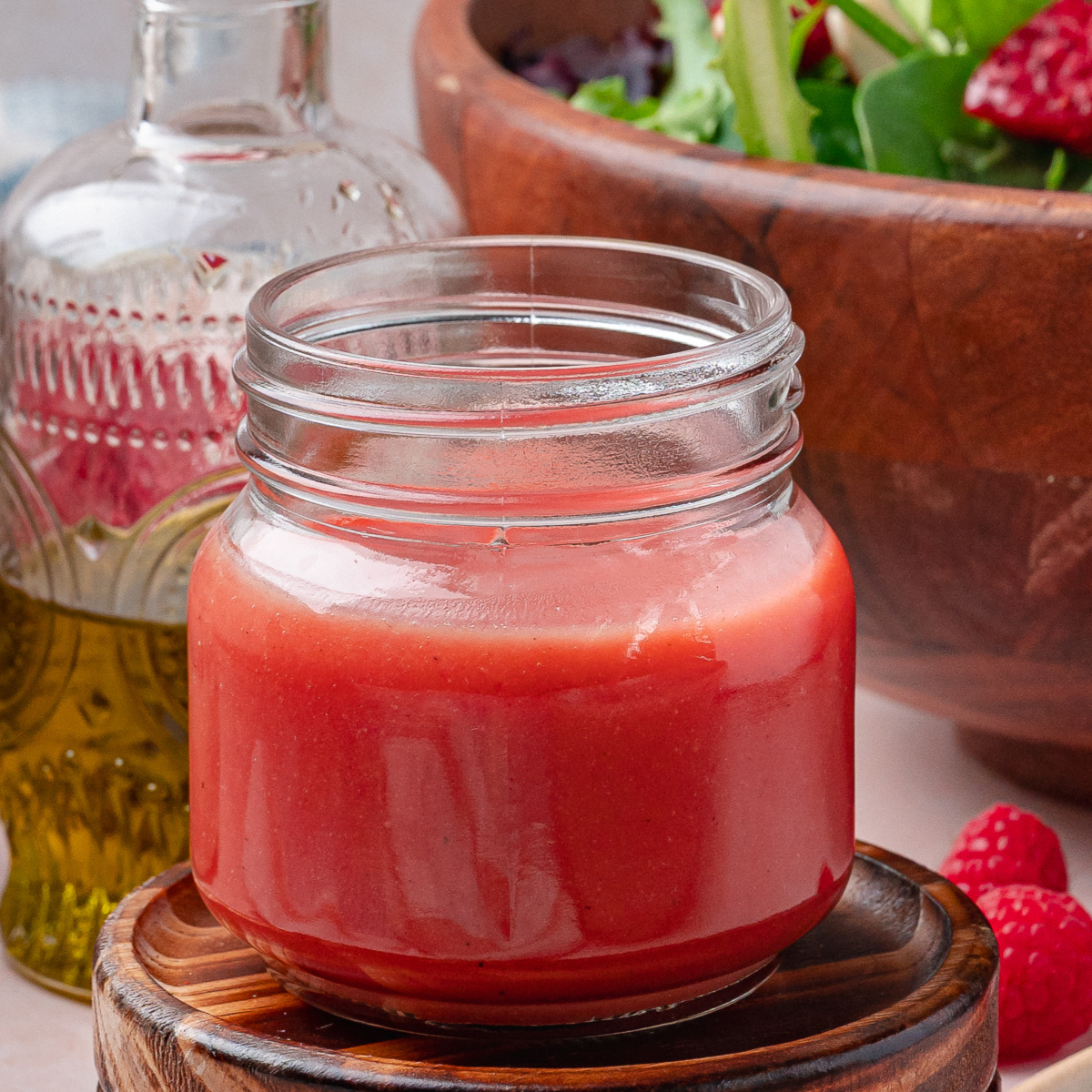 A small jar of homemade raspberry dressing resting on a wooden coaster with a salad bowl blurred in the background.