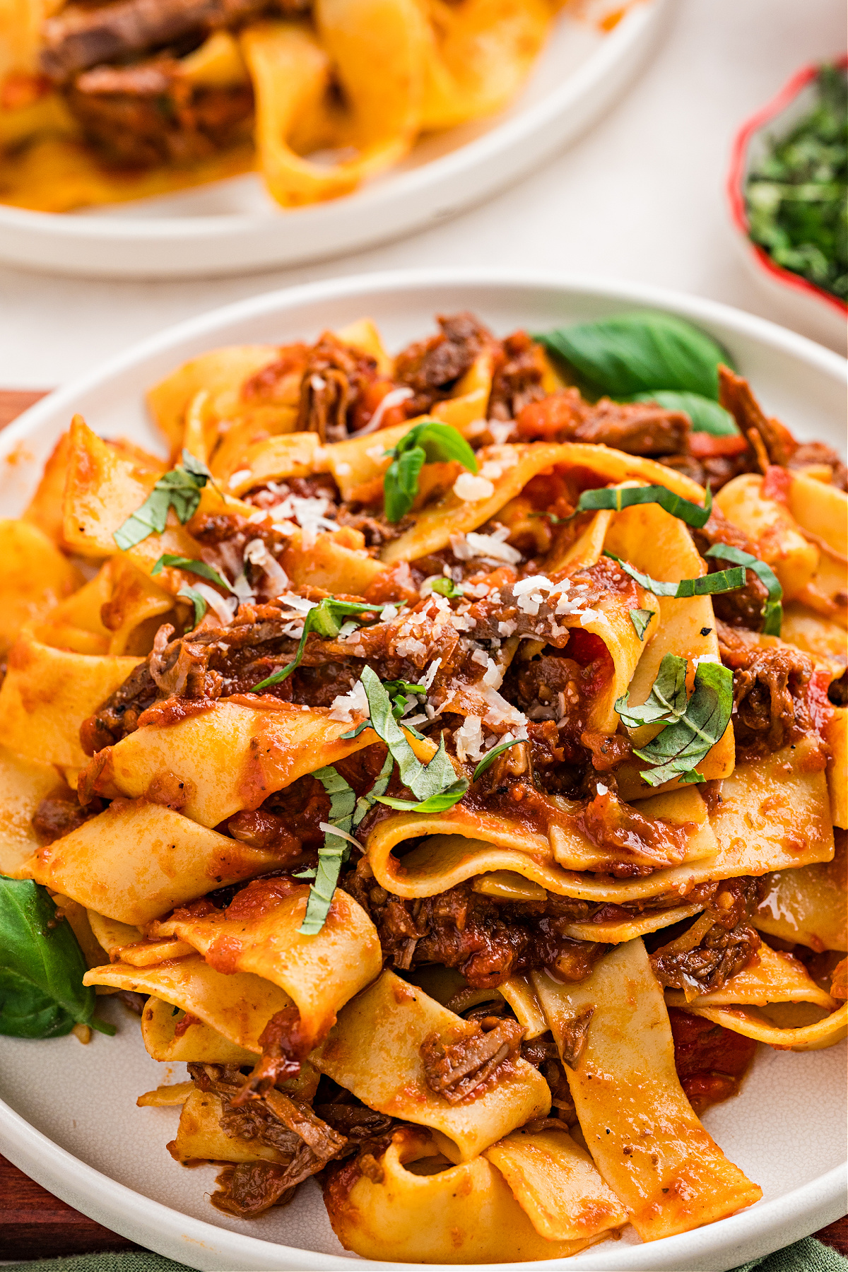 A bowl of pappardelle coated in slow-braised beef ragù with basil and parmesan. A close-up plate of slow-braised beef ragù with pappardelle, showing wide ribbons of pasta tangled with tender shredded beef, tomato-rich sauce, fresh basil ribbons, and a light grating of parmesan.
