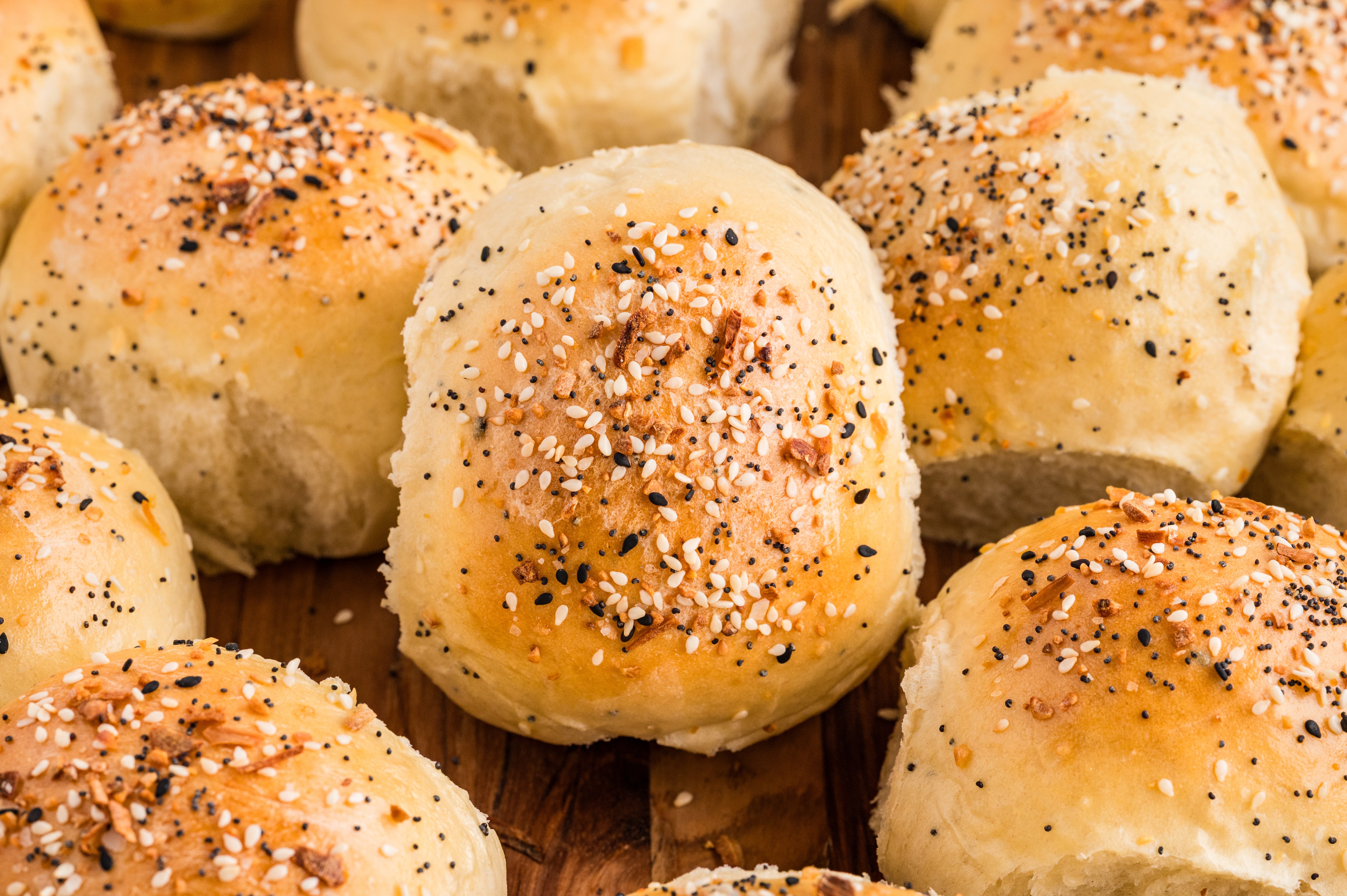 Freshly baked savory rolls topped with everything seasoning, photographed overhead on a wooden cutting board. Golden baked rolls arranged on a wooden board, topped with sesame seeds, poppy seeds, garlic, and onion, with one roll split open to show the soft interior.