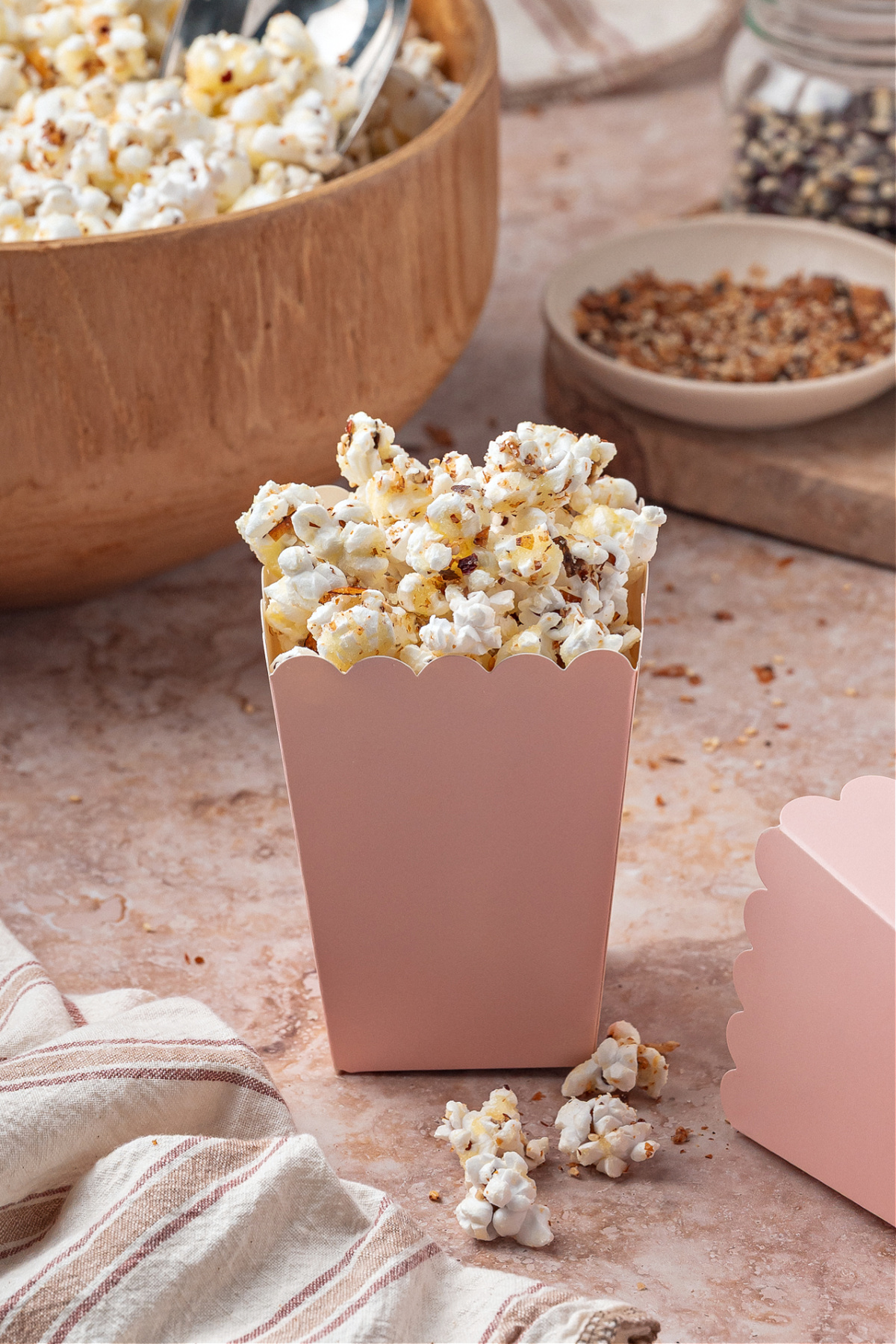 A pink carton packed with sweet and spicy furikake-seasoned popcorn. A pink snack box filled with sweet and spicy seasoned popcorn sits on a stone surface with a wooden bowl of popcorn in the background.
